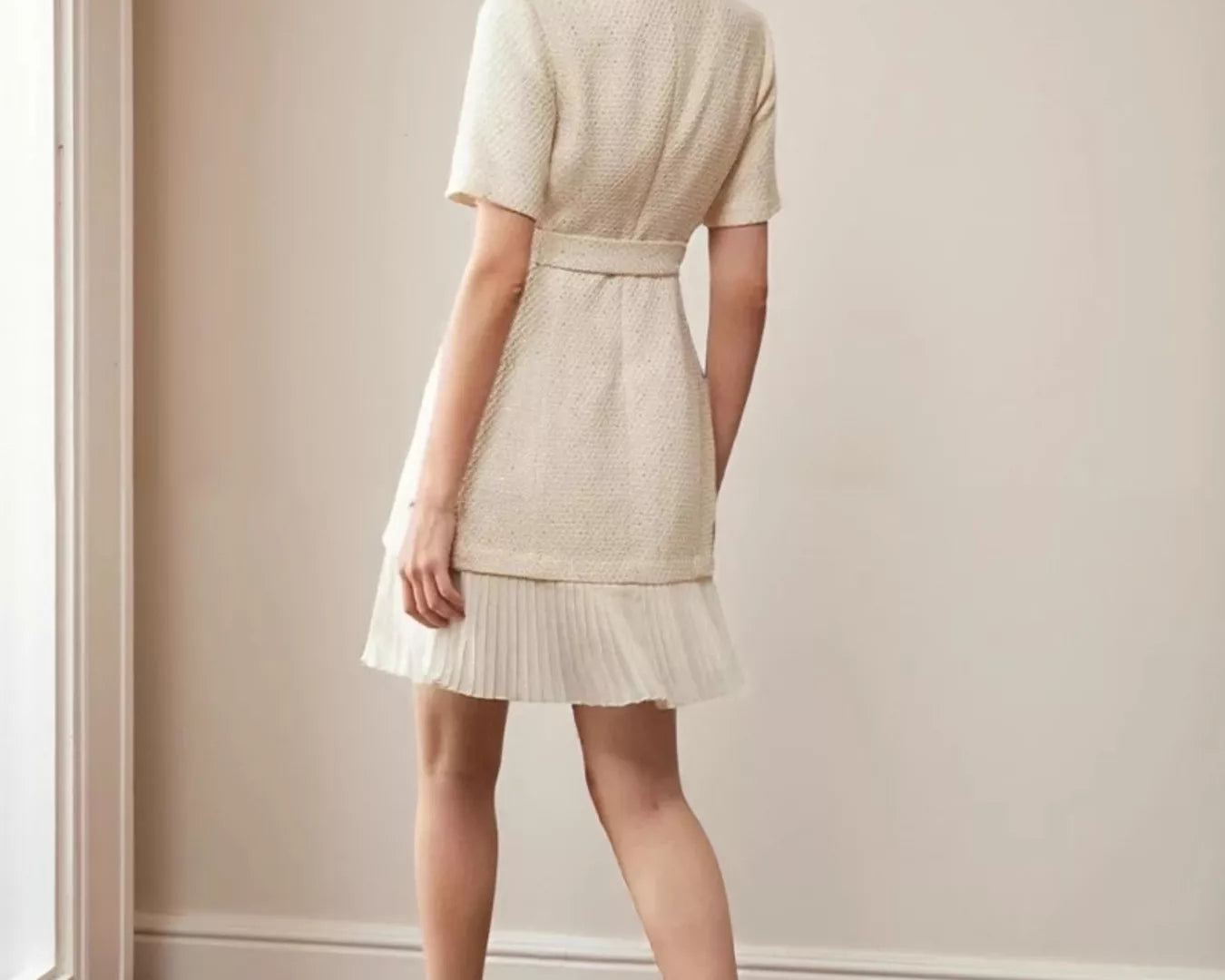 NEWYORKLISTED STYLE COLLECTION: Model wearing a beige dress standing in a room with wooden flooring and a neutral wall.
