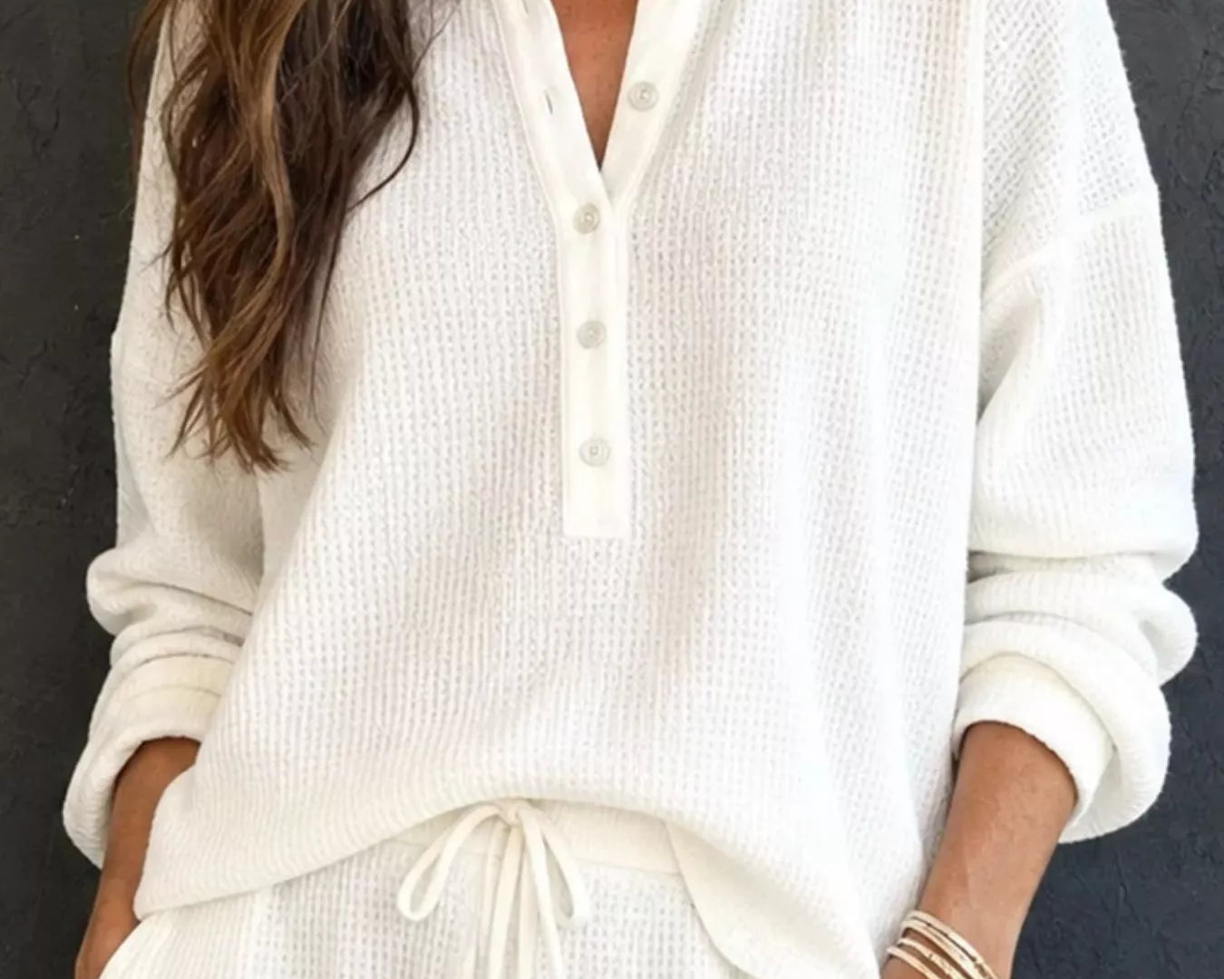 NEWYORKLISTED STYLE & COMFORT COLLECTION: A woman wearing a cream-colored waffle-knit long-sleeve henley top and matching drawstring pants, standing against a dark textured wall.