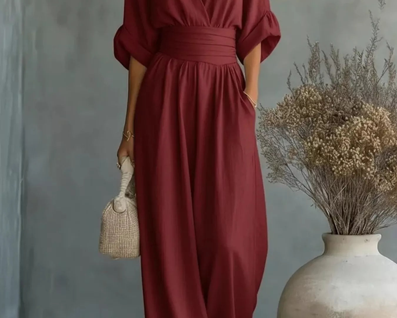 NEWYORKLISTED STYLE COLLECTION: Woman wearing a burgundy jumpsuit holding a woven bag against a gray wall with a vase of dried flowers.