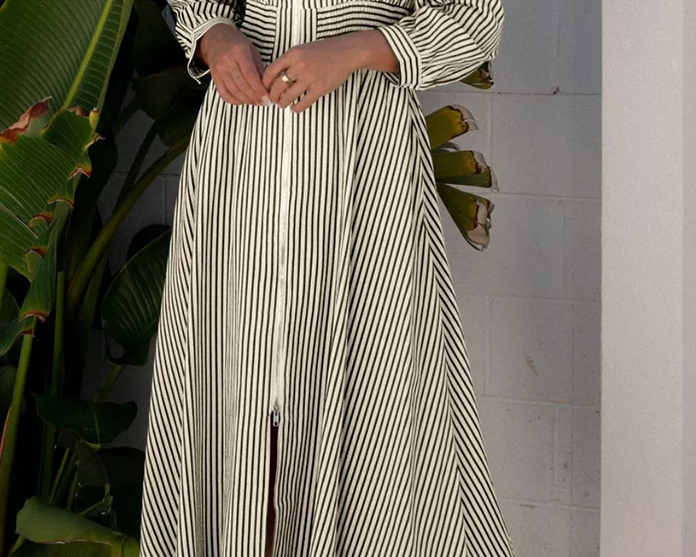 NEWYORKLISTED STYLE COLLECTION: Woman wearing a striped dress standing outdoors with plants and a white wall in the background