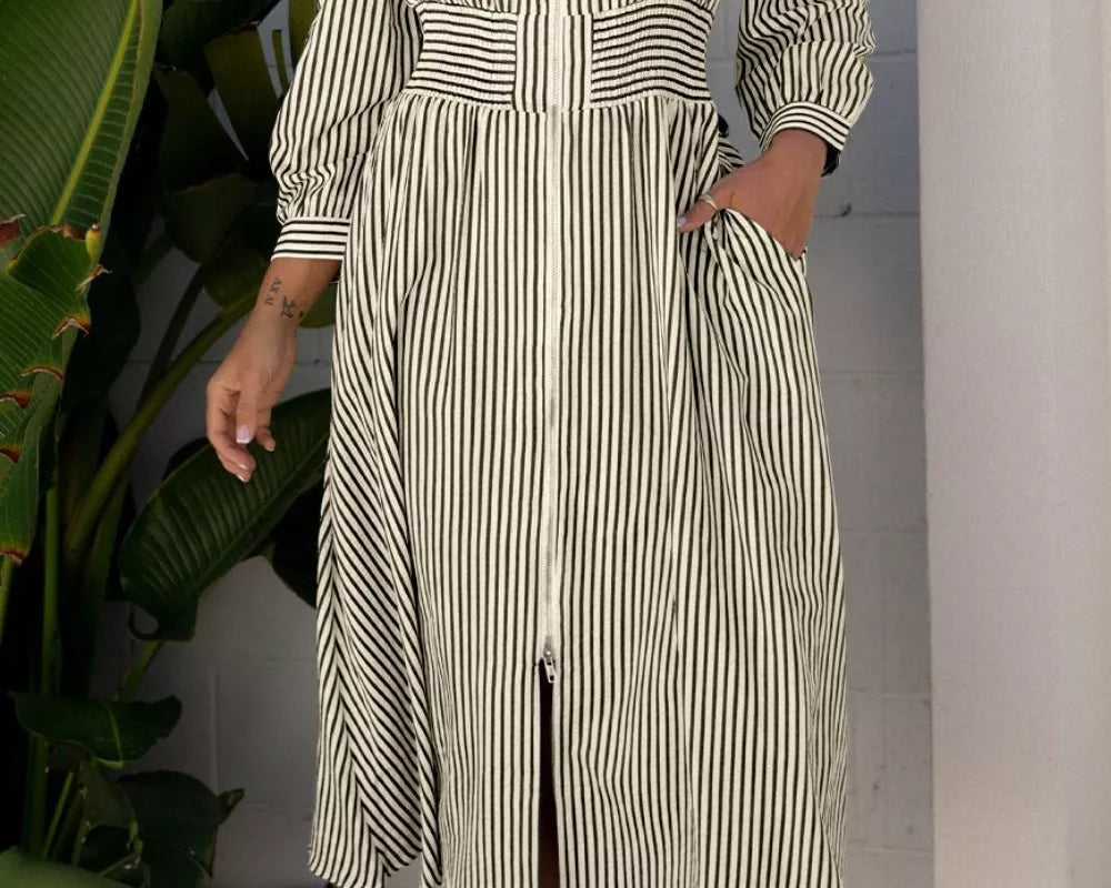 NEWYORKLISTED STYLE COLLECTION: Woman wearing a striped dress standing outdoors with plants and a white wall in the background