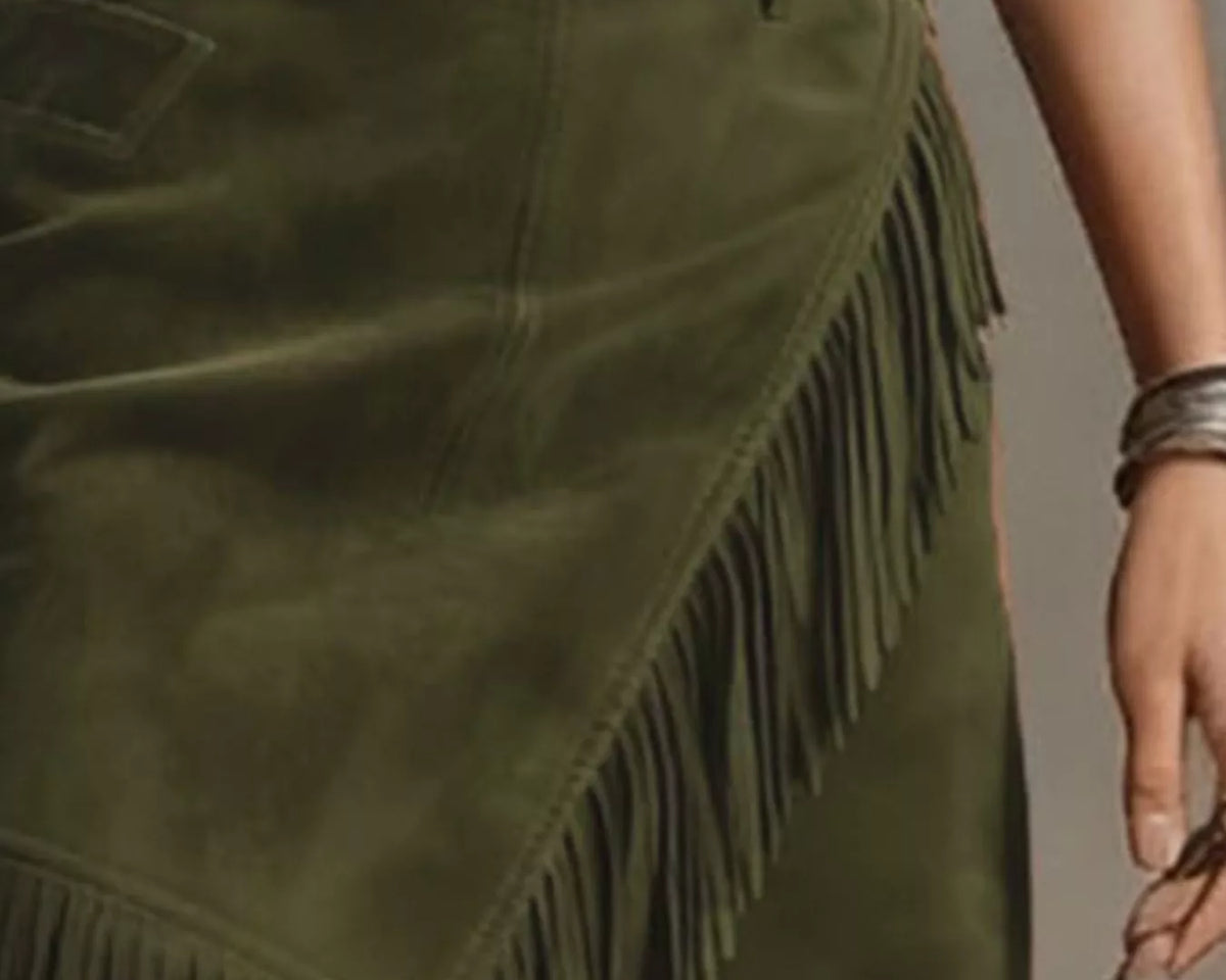 NEWYORKLISTED STYLE COLLECTION: An olive green suede skirt with long fringe trim, shown on a model. The skirt features an asymmetrical design with a side pocket and a belt loop.