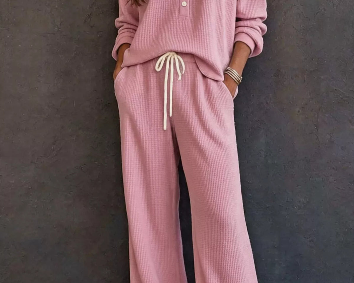 NEWYORKLISTED STYLE & COMFORT COLLECTION: A woman wearing a matching dusty pink lounge set consisting of a long-sleeve button-front top and wide-leg pants with a cream drawstring, standing against a dark textured wall.