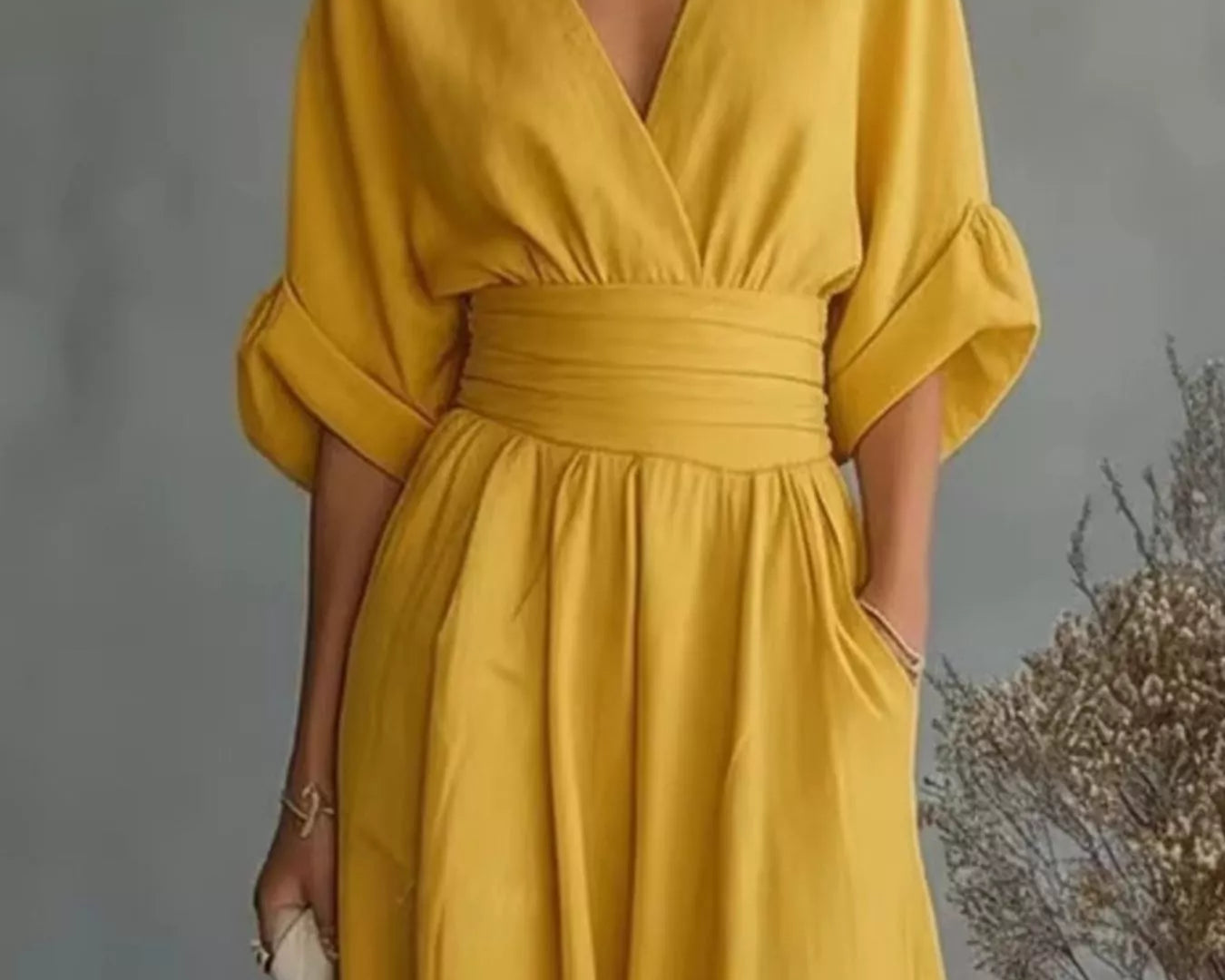 NEWYORKLISTED STYLE COLLECTION: A woman wearing a mustard yellow midi dress with a V-neckline, wide elbow-length sleeves, a matching fabric waist belt, and side pockets, holding a woven cream handbag against a gray studio backdrop with dried floral decor.