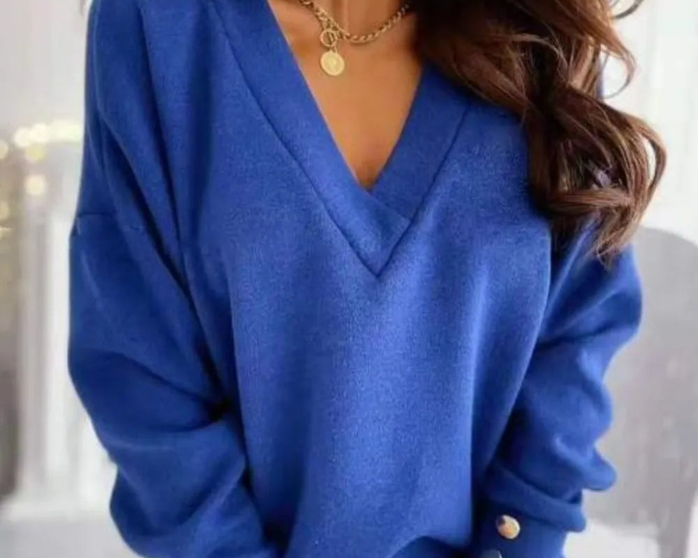 NEWYORKLISTED STYLE COLLECTION: A woman wearing a royal blue V-neck knit sweater with decorative gold-tone buttons on the cuffs, paired with light beige pants and a gold necklace.