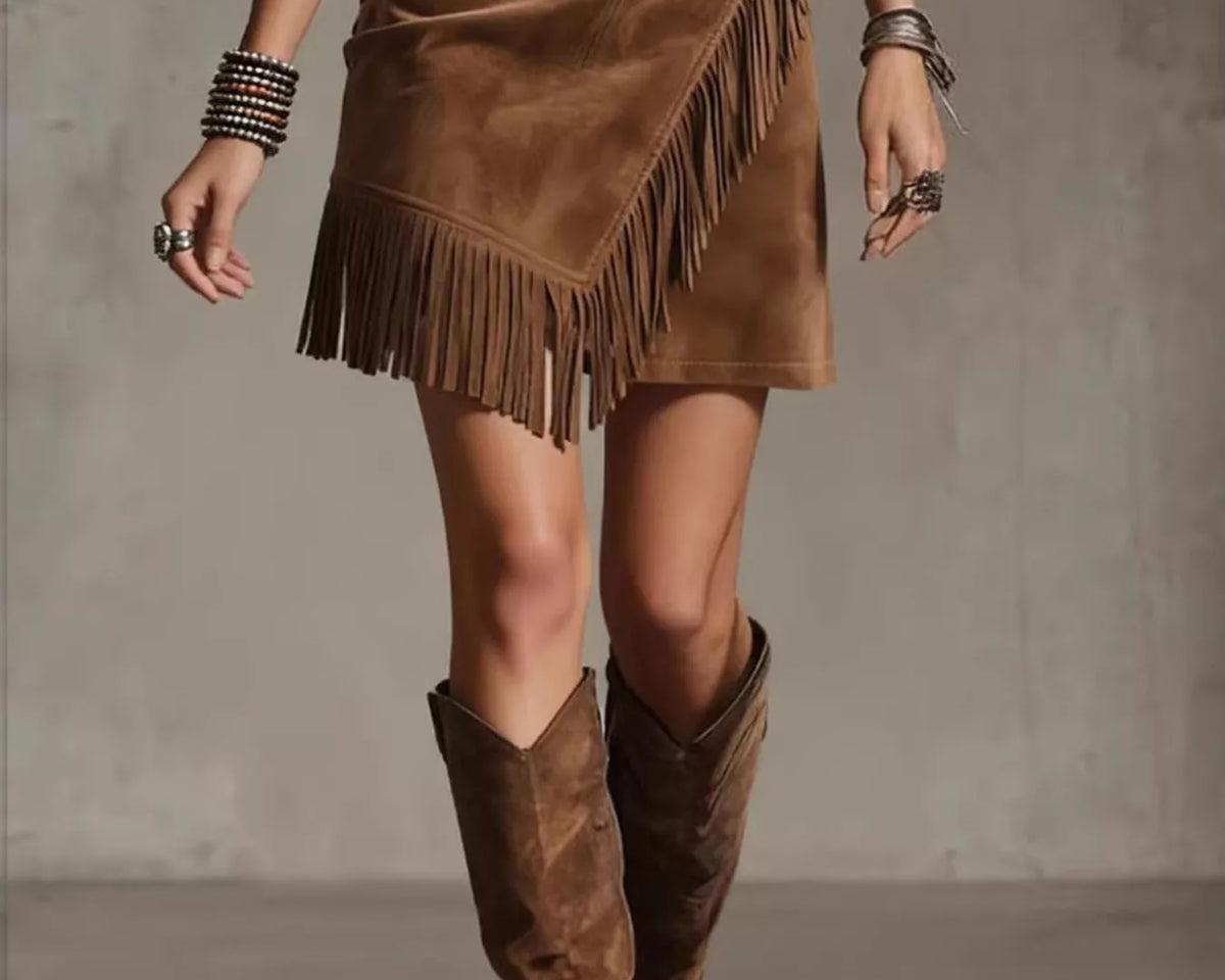 NEWYORKLISTED STYLE COLLECTION: A woman wearing a brown suede fringe mini skirt with an asymmetrical wrap design, paired with matching brown suede knee-high boots and silver bracelets, set against a textured gray studio background.