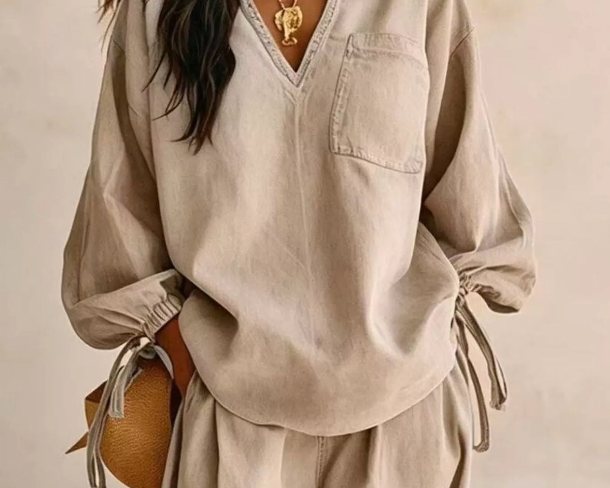 NEWYORKLISTED STYLE COLLECTION: A woman wearing a beige linen-like two-piece set with a long-sleeve V-neck top featuring a chest pocket and matching wide-leg pants, styled with gold jewelry and a straw bag against a neutral beige background.