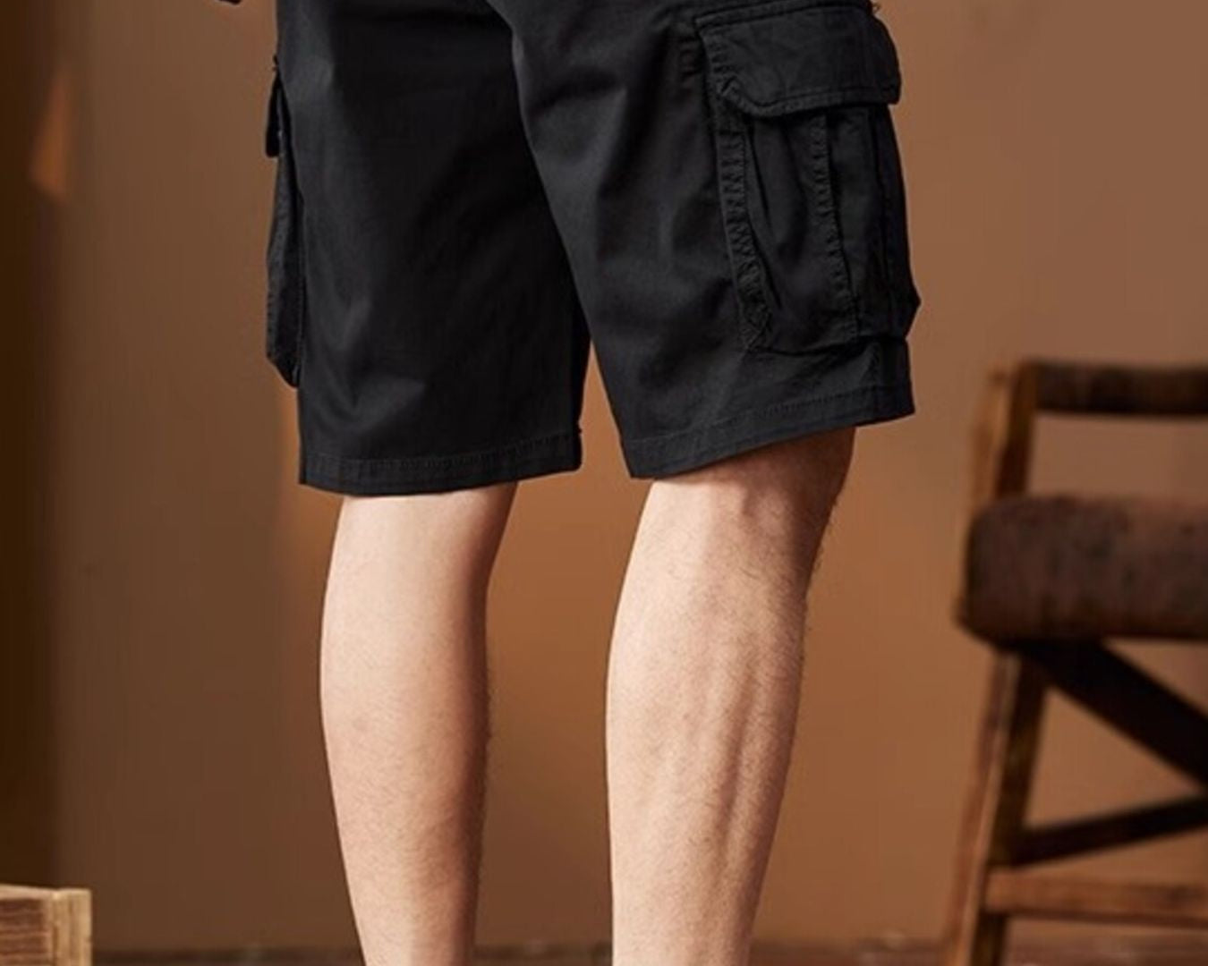 NEWYORKLISTED ATLAS MEN's COLLECTION: Person wearing black cargo shorts and tan boots in a room with a wooden floor and chair.