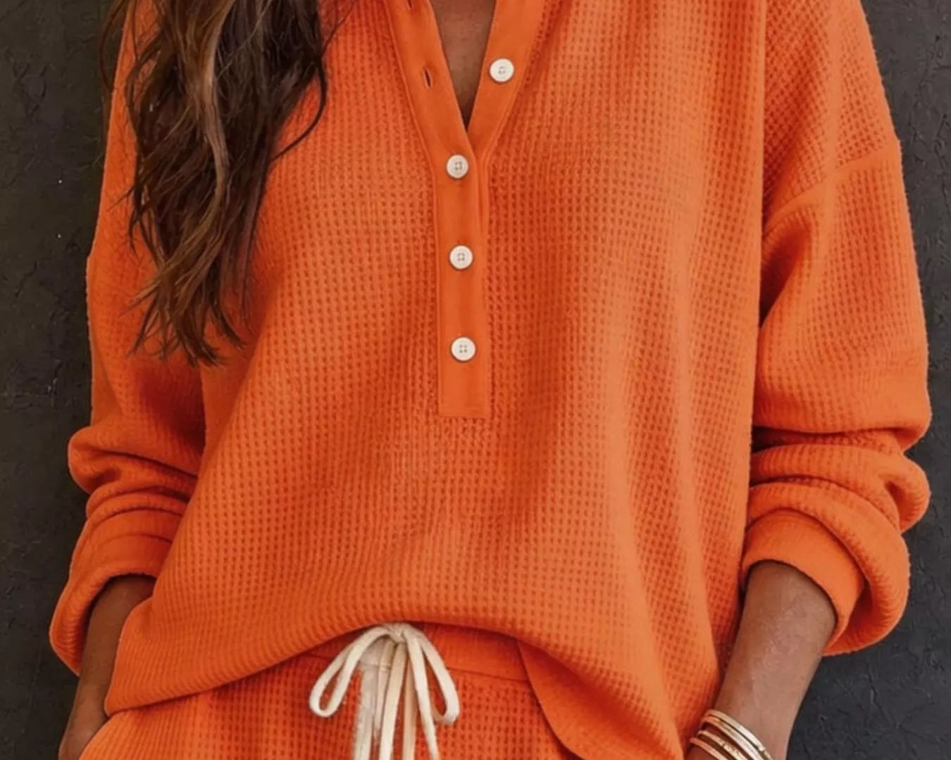NEWYORKLISTED STYLE & COMFORT COLLECTION: A woman wearing a bright orange waffle-knit two-piece loungewear set, featuring a long-sleeve henley top with white buttons and matching drawstring pants, set against a dark textured wall.