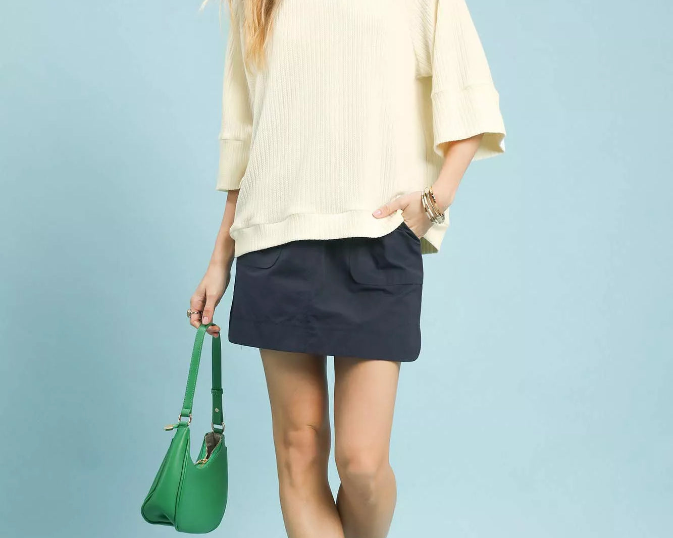 NEWYORKLISTED STYLE COLLECTION: Woman wearing a beige top and navy skirt with a green handbag on a light blue background