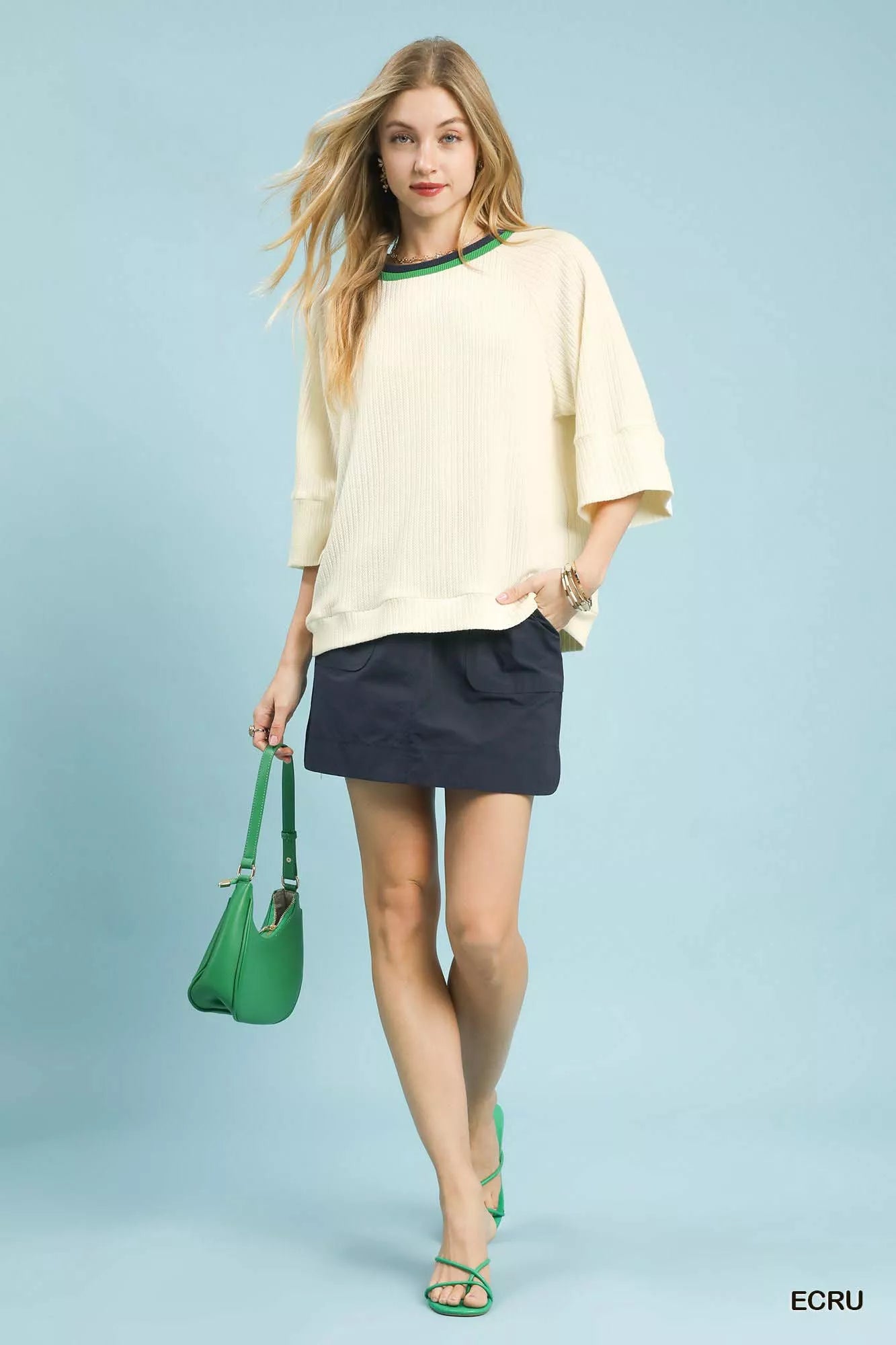 NEWYORKLISTED STYLE COLLECTION: Woman wearing a beige top and navy skirt with a green handbag on a light blue background