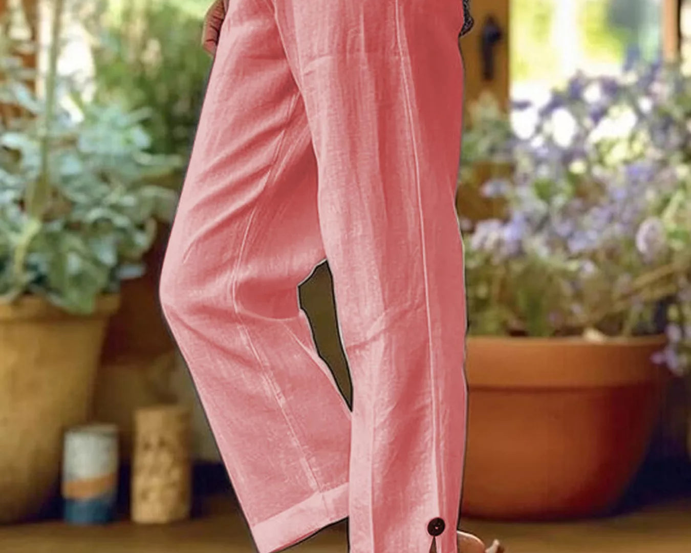 NEWYORKLISTED STYLE COLLECTION: A person wearing loose pink cropped pants with a button detail at the hem, paired with a patterned top and sandals, standing in a sunlit indoor setting with potted plants in the background.