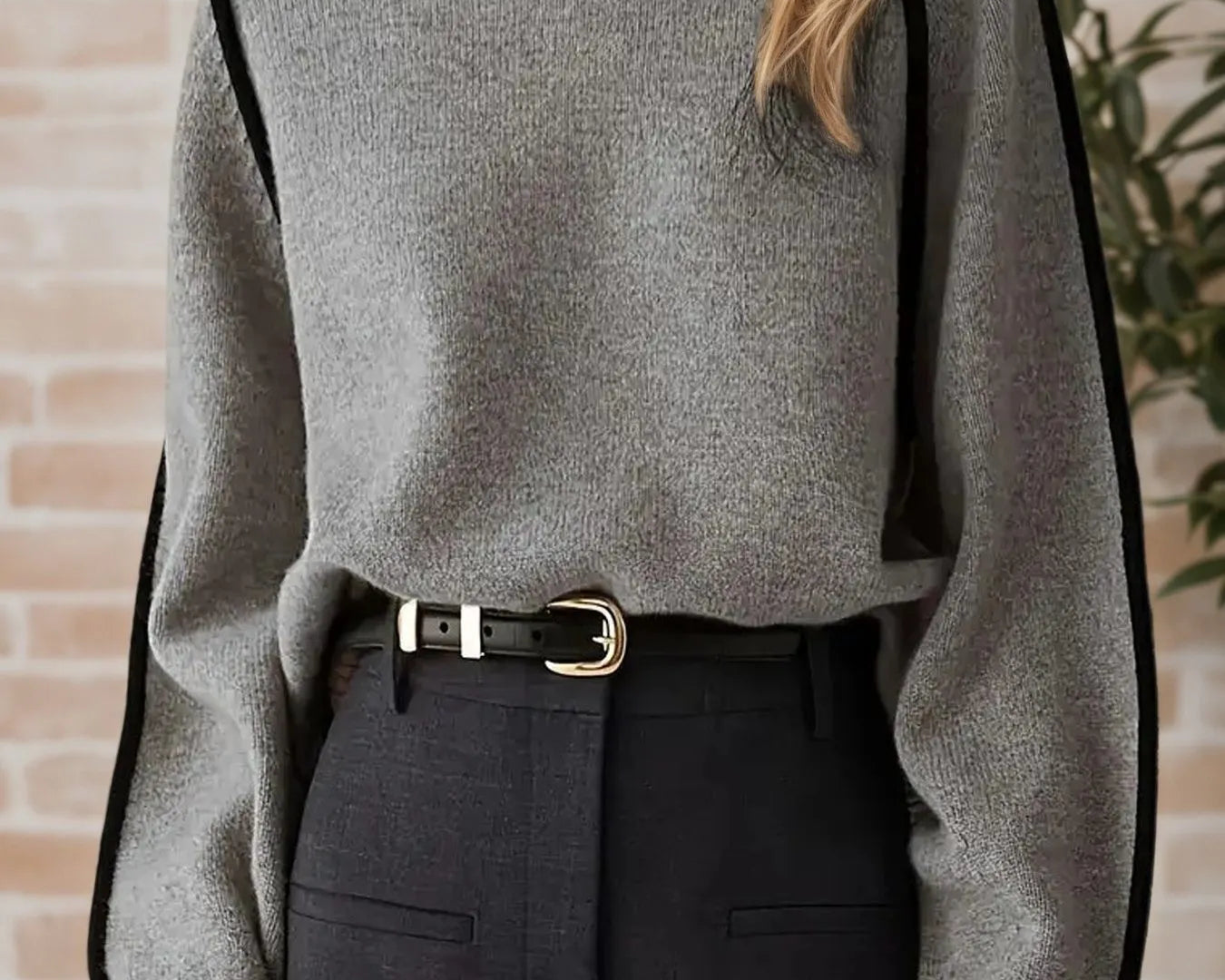 NEWYORKLISTE STYLE COLELCTION: Person wearing a gray sweater with black accents and black pants against a brick wall.