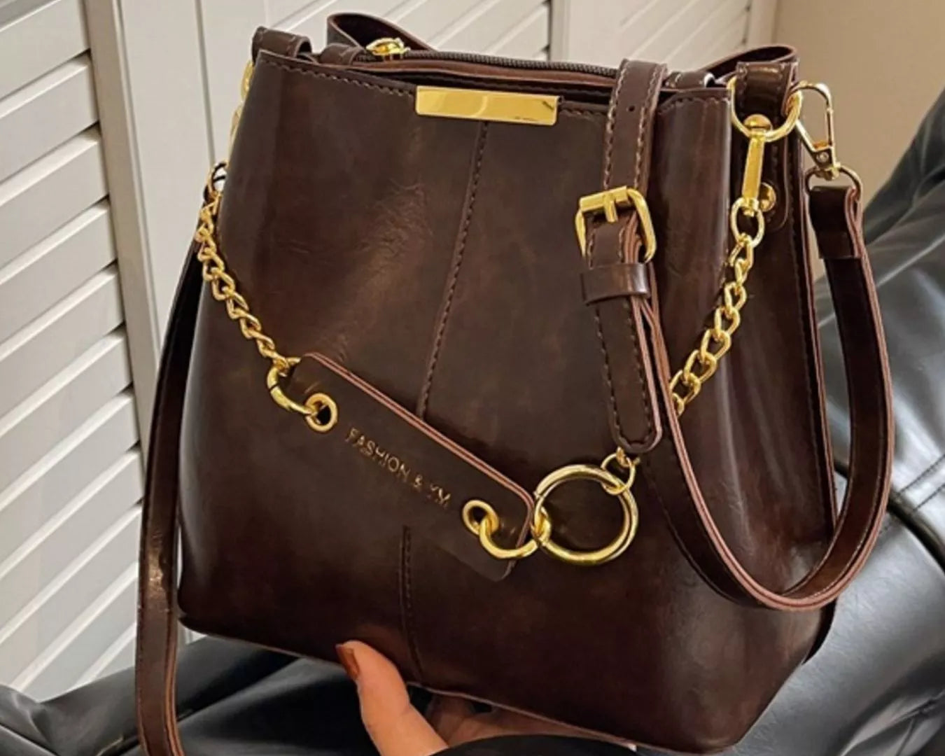 Brown handbag with gold accents held by a person against a neutral background New York Listed Style Collection