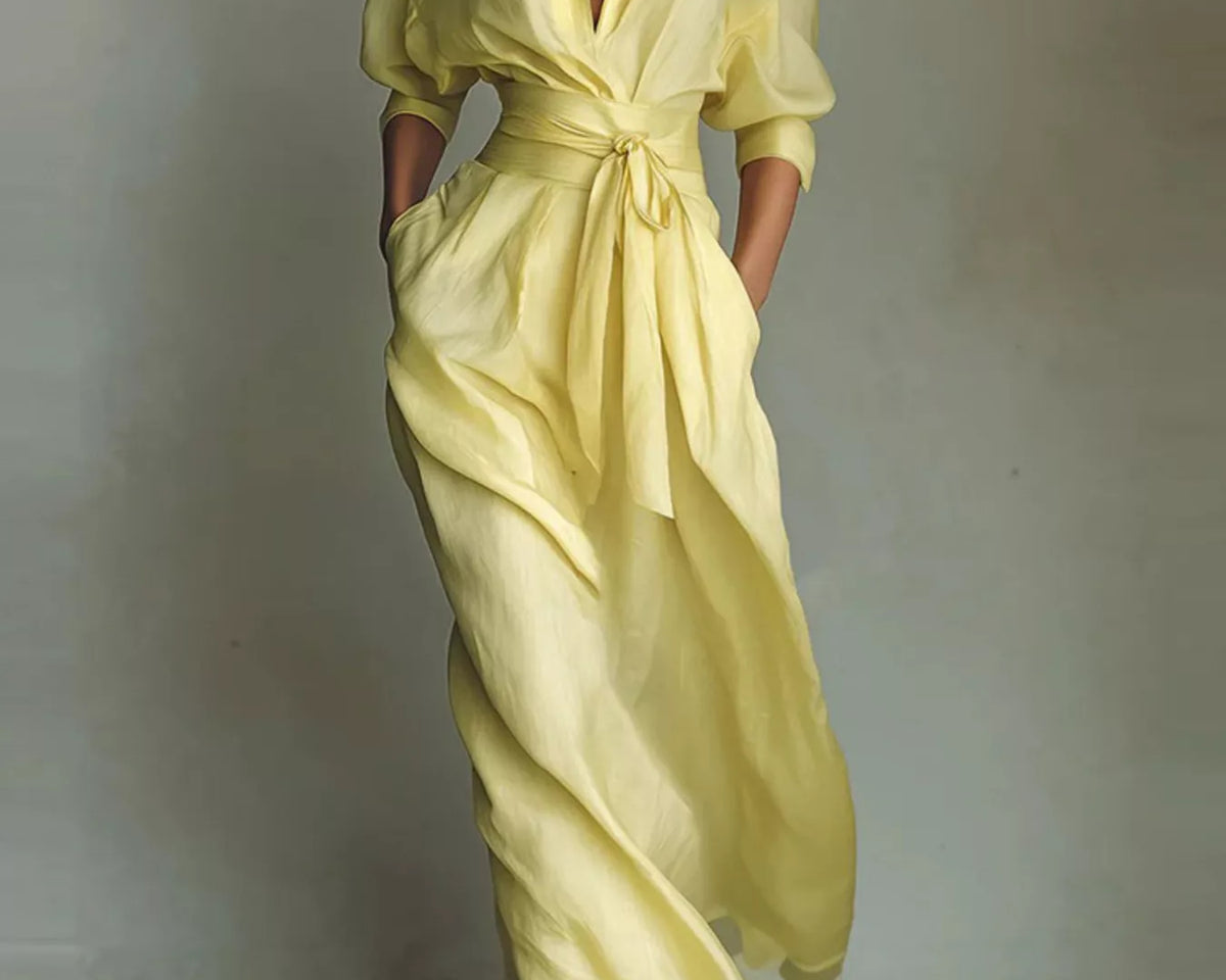 NEWYORKLISTED STYLE COLLECTION: A woman models a flowing pale yellow wrap-style maxi dress with a deep V-neck, short sleeves, side pockets, and a self-tie waist, photographed against a neutral textured wall.