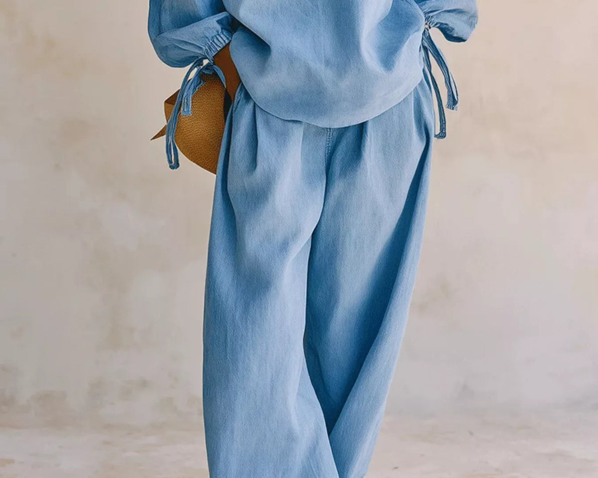 NEWYORKLISTED STYLE COLLECTION: A person wearing a light blue matching lounge set with a loose long-sleeve top and wide-leg pants, standing against a neutral beige wall.