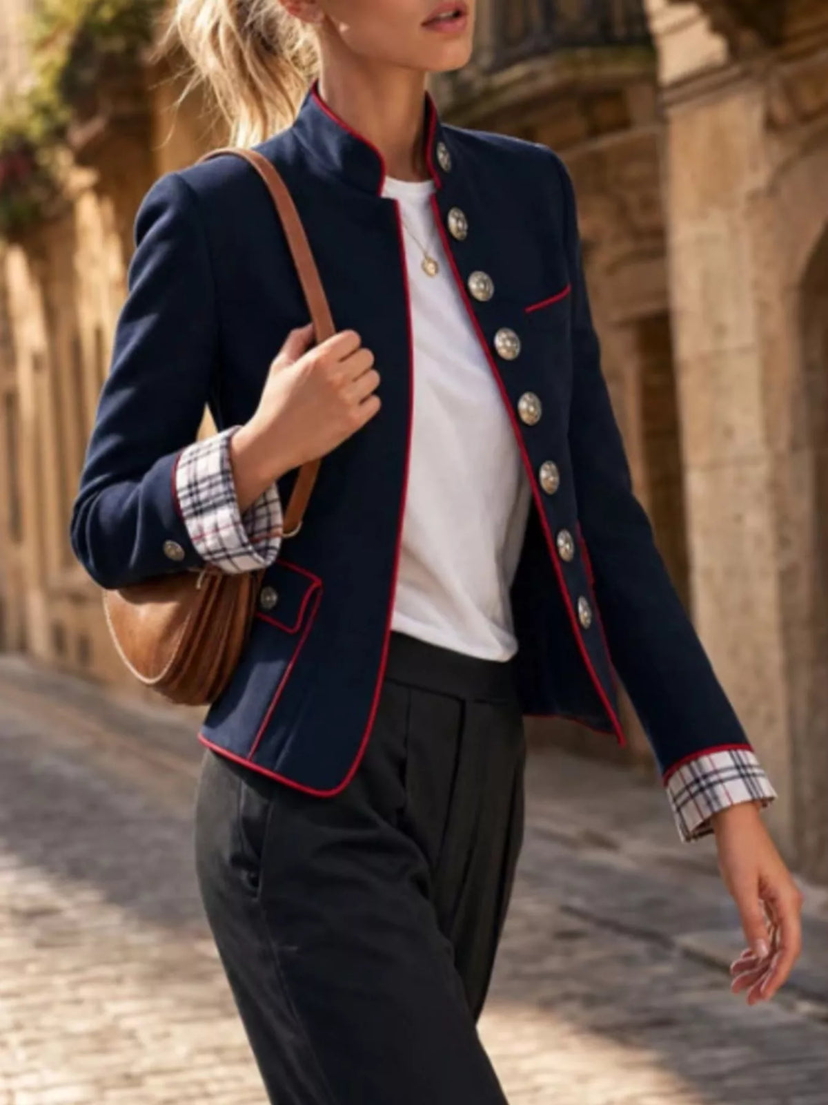 NEWYORKLISTED STYLE COLLECTION: A woman wearing a navy blue military-style blazer with red piping, silver buttons, and a plaid cuff, paired with a white top and dark trousers, carrying a brown shoulder bag in an outdoor stone alleyway.