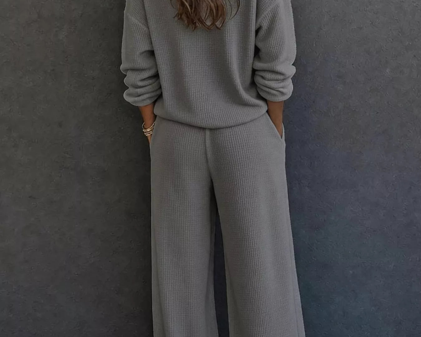NEWYORKLISTED STYLE & COMFORT COLLECTION: A woman viewed from behind wearing a matching gray ribbed knit lounge set consisting of a long-sleeve top and wide-leg pants, standing against a dark gray textured wall.