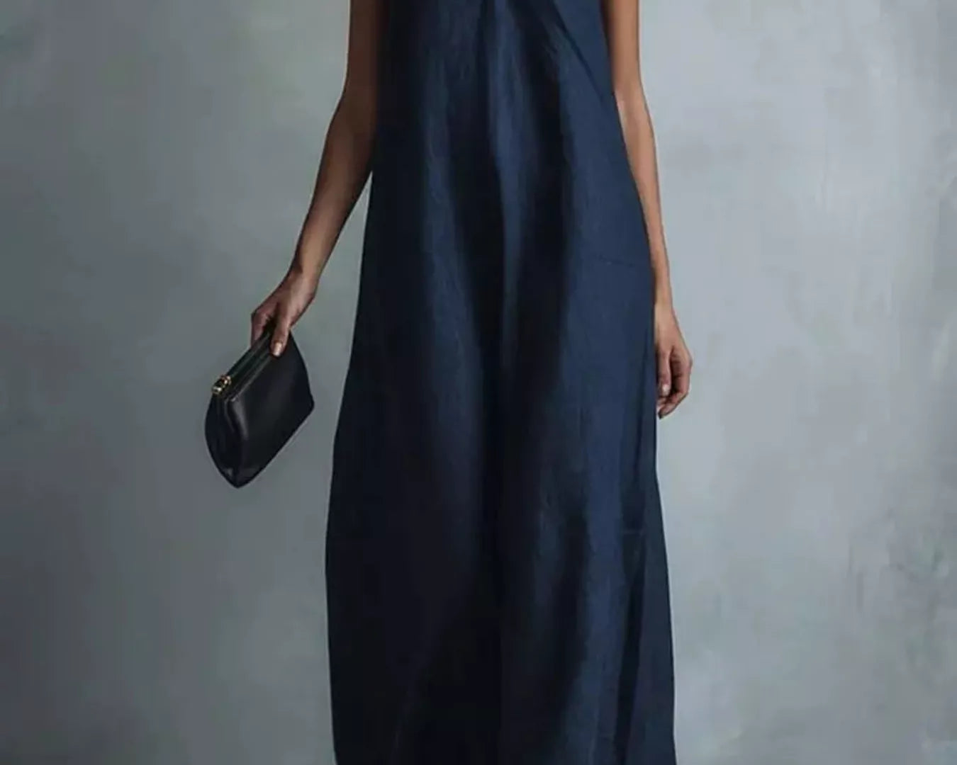 NEWYORKLISTED STYLE COLLECTION: A woman models a sleeveless navy blue maxi dress with a V-neckline, holding a small black clutch against a textured gray studio background.
