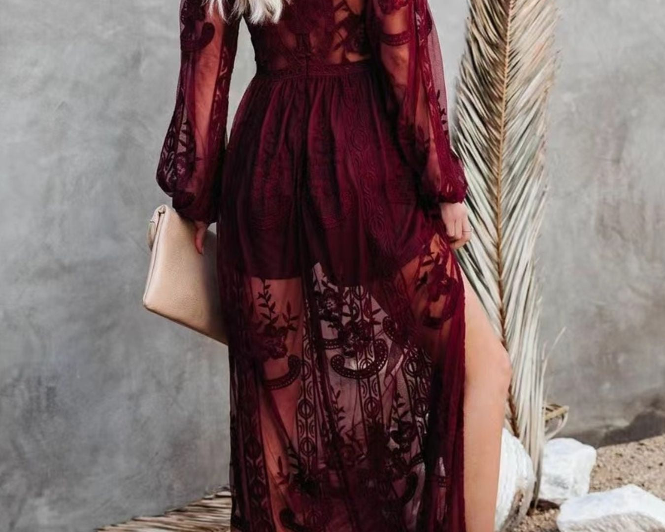 NEWYORKLISTED STYLE COLLECTION: Woman wearing a burgundy lace dress with long sleeves and a slit, standing against a textured wall.