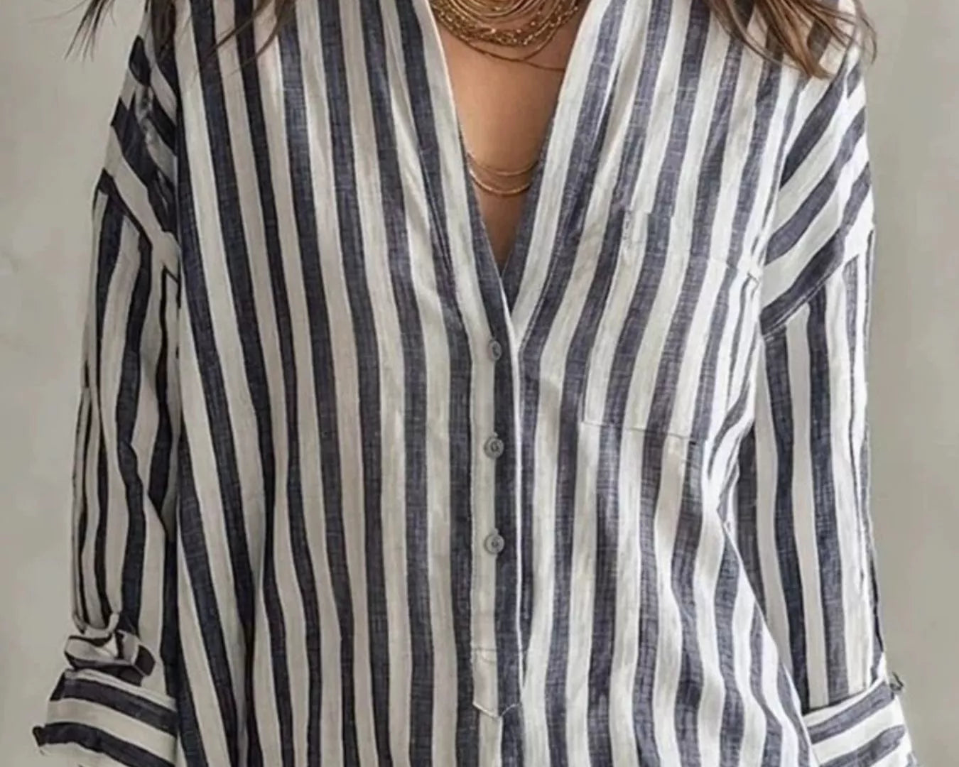 NEWYORKLISTED STYLE COLLECTION: A woman wearing a loose-fitting linen shirt dress with vertical navy and off-white stripes, styled with layered gold necklaces and earrings.