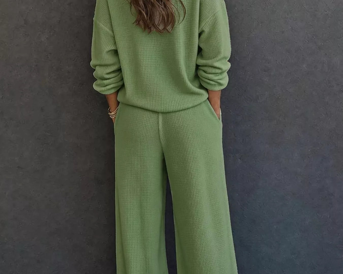NEWYORKLISTED STYLE & COMFORT COLLECTION: A woman viewed from behind wearing a matching sage green knit lounge set consisting of a long-sleeve top and wide-leg pants, standing against a dark gray textured wall.