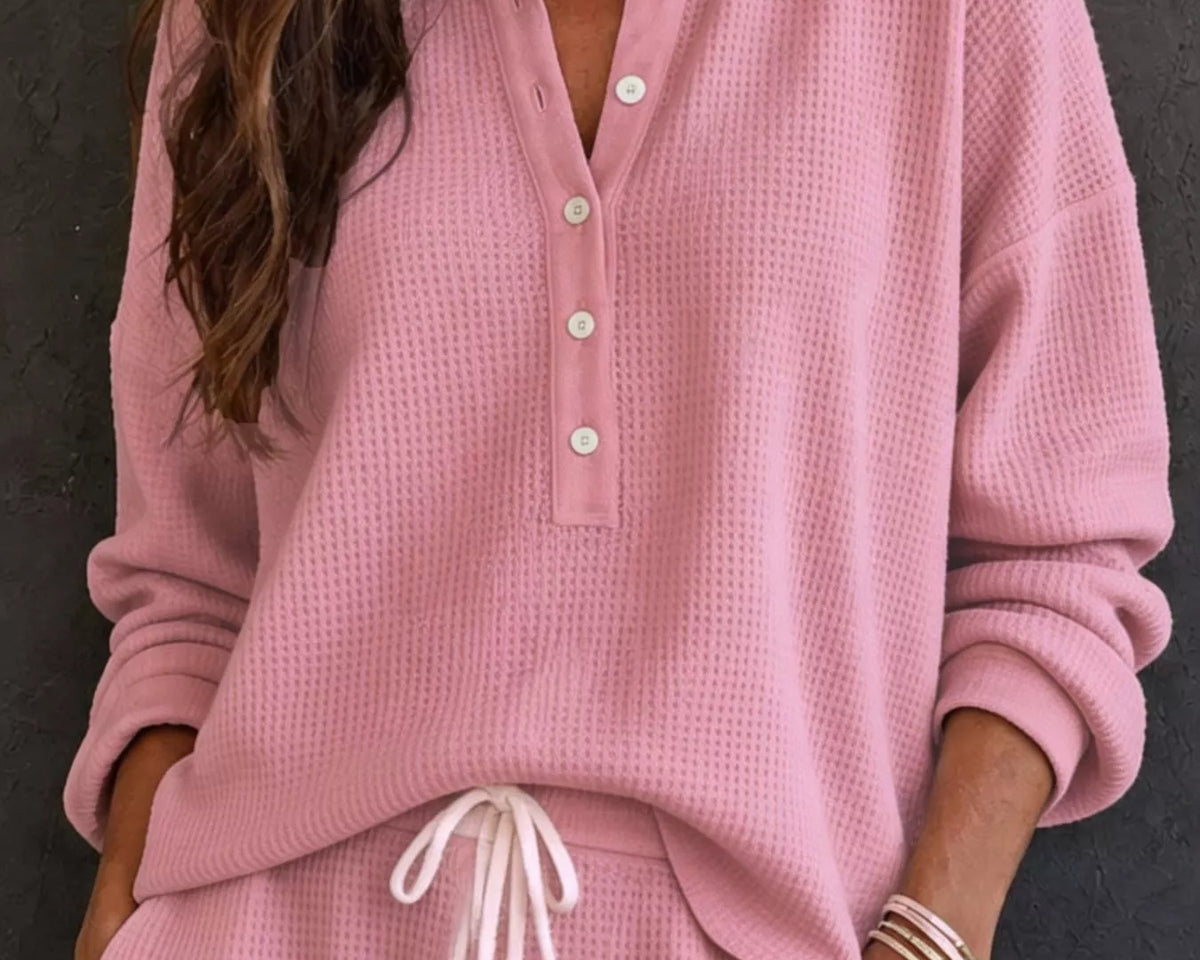NEWYORKLISTED STYLE & COMFORT COLLECTION: A woman wearing a matching pink waffle-knit lounge set with a henley top and drawstring pants, photographed against a dark textured wall.
