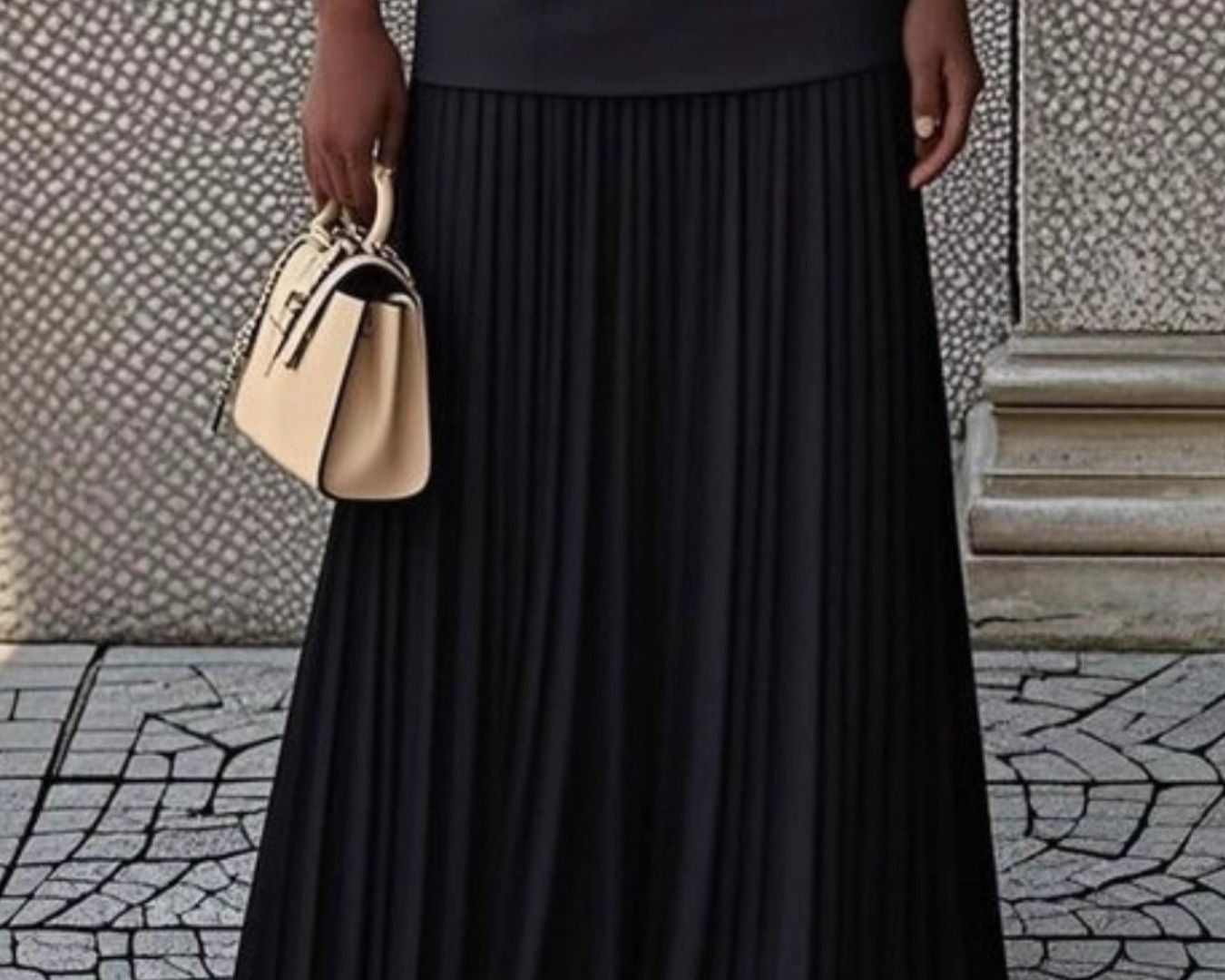 NEWYORKLISTED STYLE COLLECTION: Person wearing a black pleated skirt holding a beige handbag on a stone pavement.