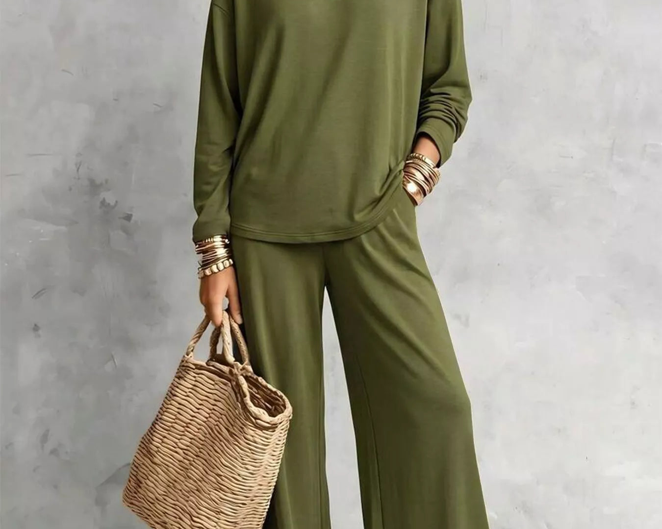 NEWYORKLISTED STYLE COLLECTION: A woman models an olive green two-piece lounge set consisting of a long-sleeve top and wide-leg pants, styled with layered gold necklaces, bracelets, and a straw tote bag against a textured gray wall.