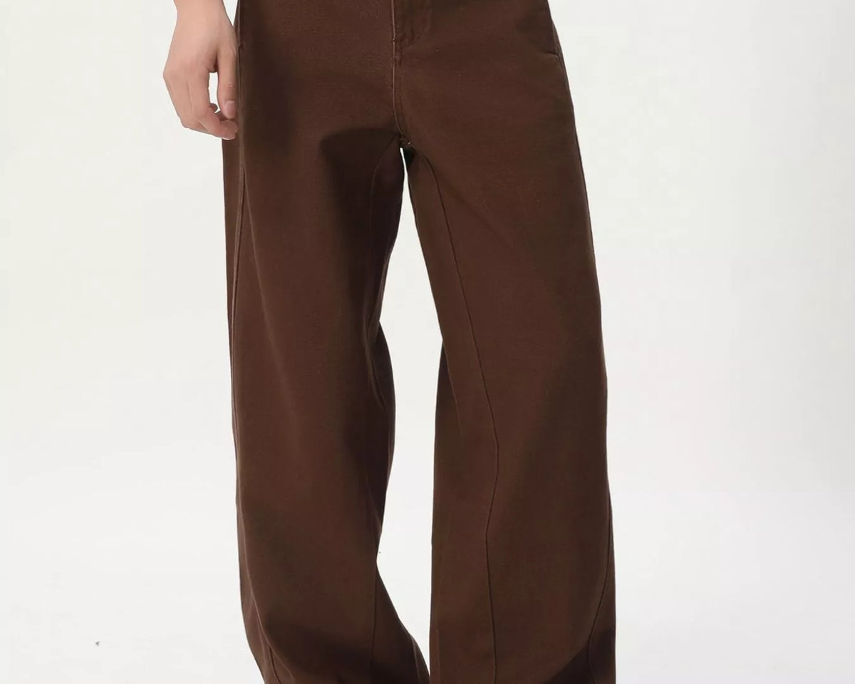 NEWYORKLISTED ATLAS MEN'S COLLECTION: A person wearing loose brown wide-leg pants with a white graphic t-shirt and beige suede shoes, standing against a plain white studio background.