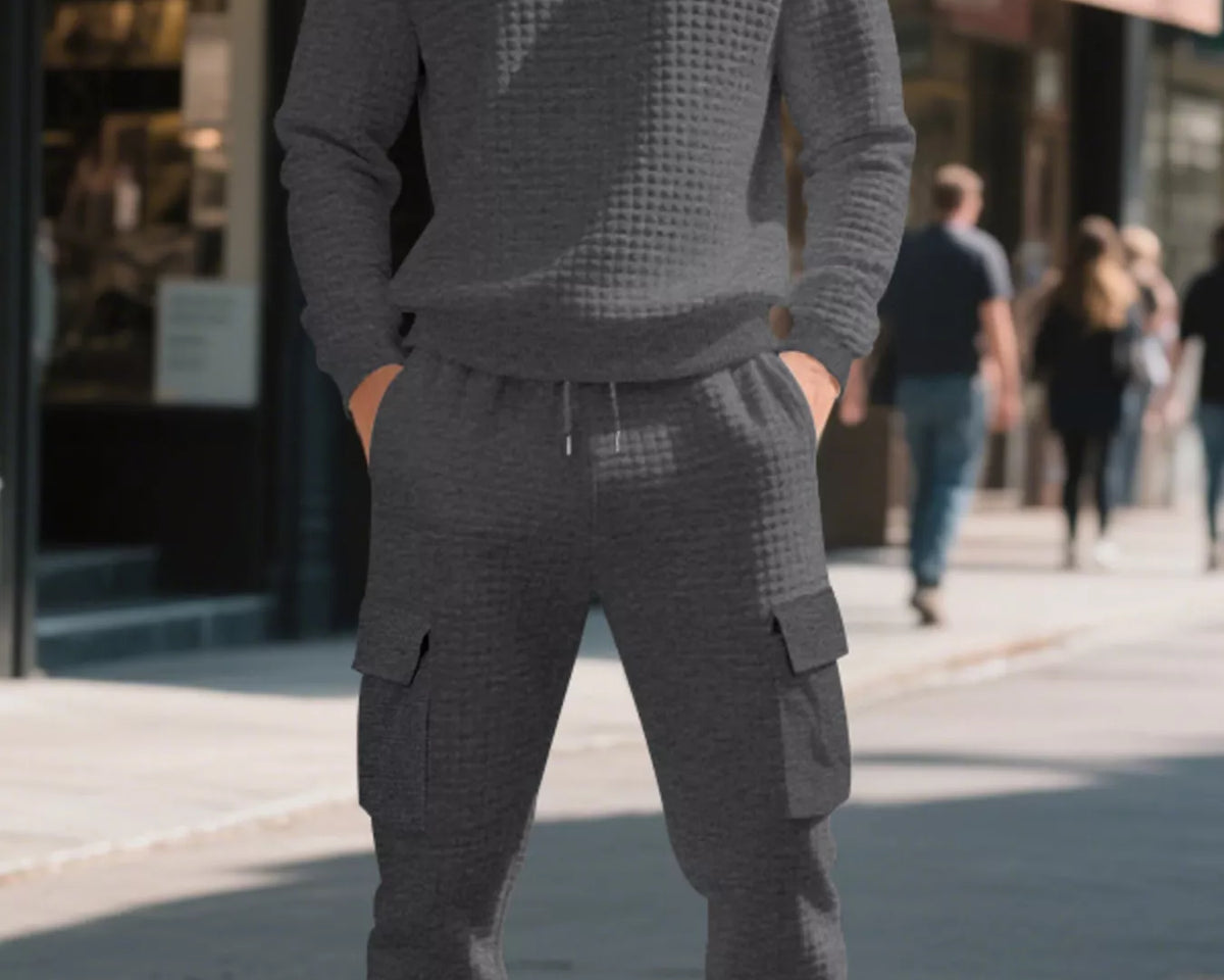 NEWYORKLISTED ATLAS MEN'S COLLECTION: A man wearing a matching dark gray waffle-knit crewneck sweatshirt and jogger cargo pants stands on a city sidewalk with blurred storefronts and pedestrians in the background.