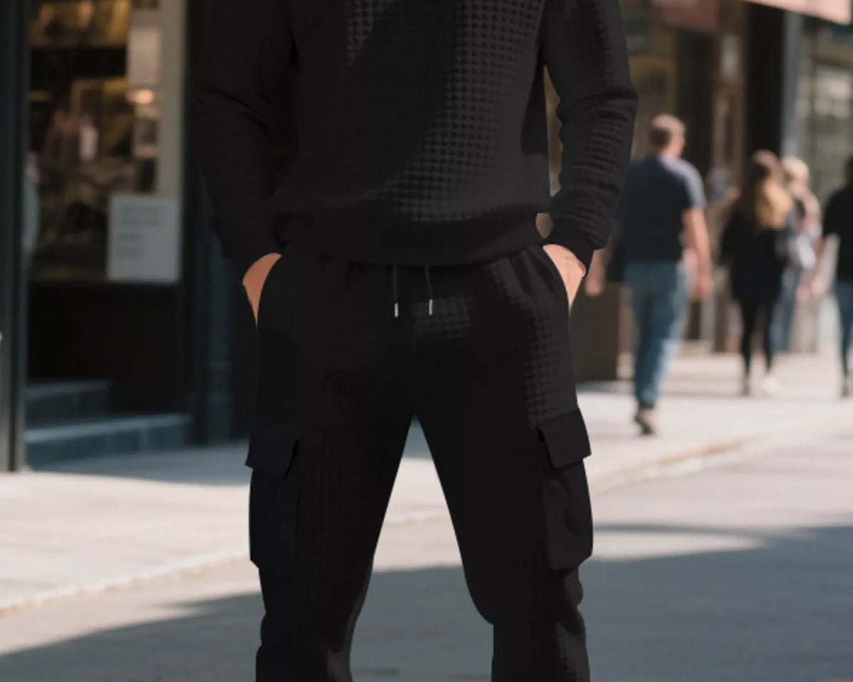 NEWYORKLISTED ATLAS MEN'S COLLECTION: A man wearing a black textured crewneck sweatshirt and matching black cargo jogger pants stands on a city sidewalk with blurred storefronts and pedestrians in the background.
