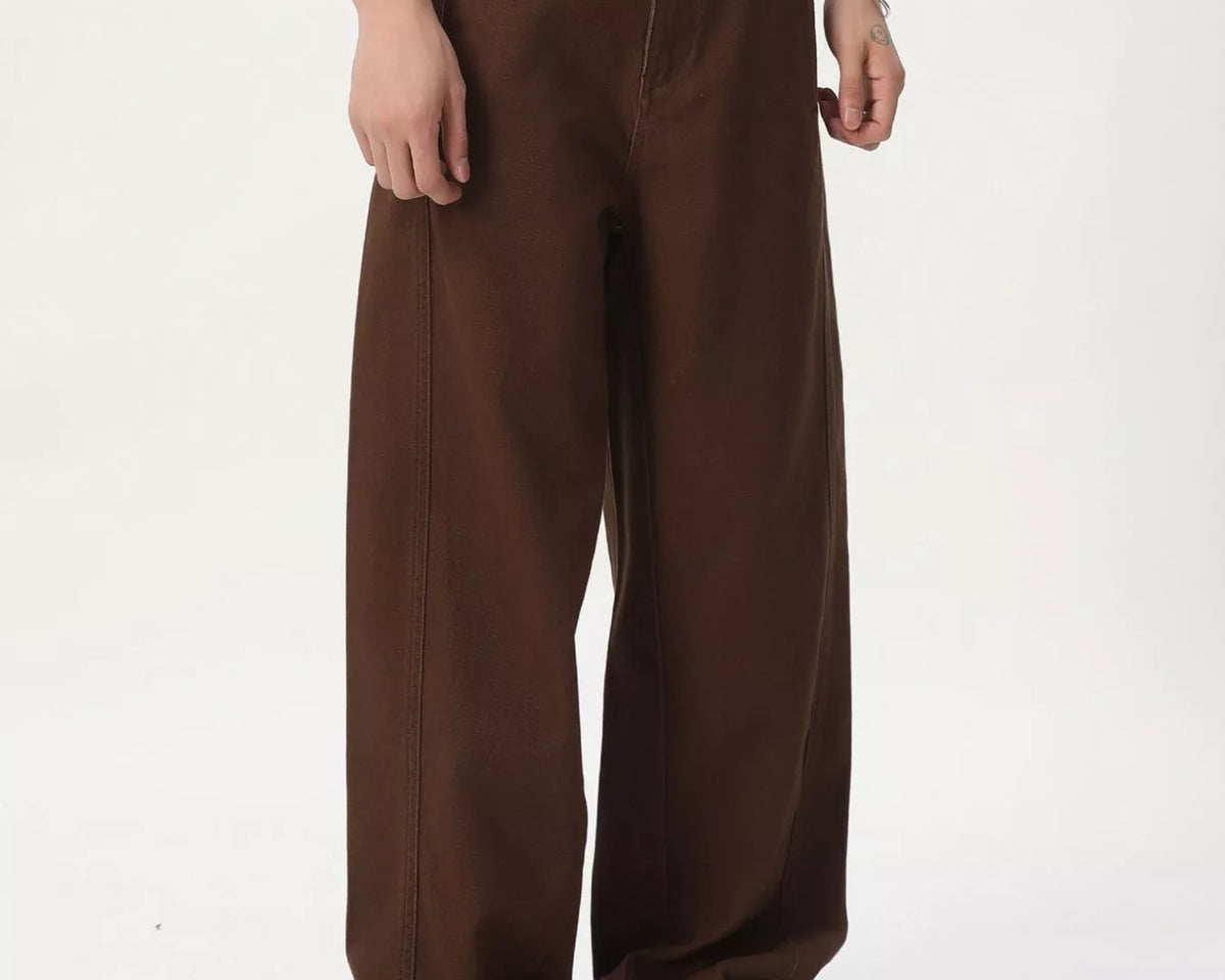 NEWYORKLISTED ATLAS MEN'S COLLECTION: A person wearing loose, wide-leg brown pants with a black belt, a white top, and tan slip-on shoes, photographed against a plain light background.