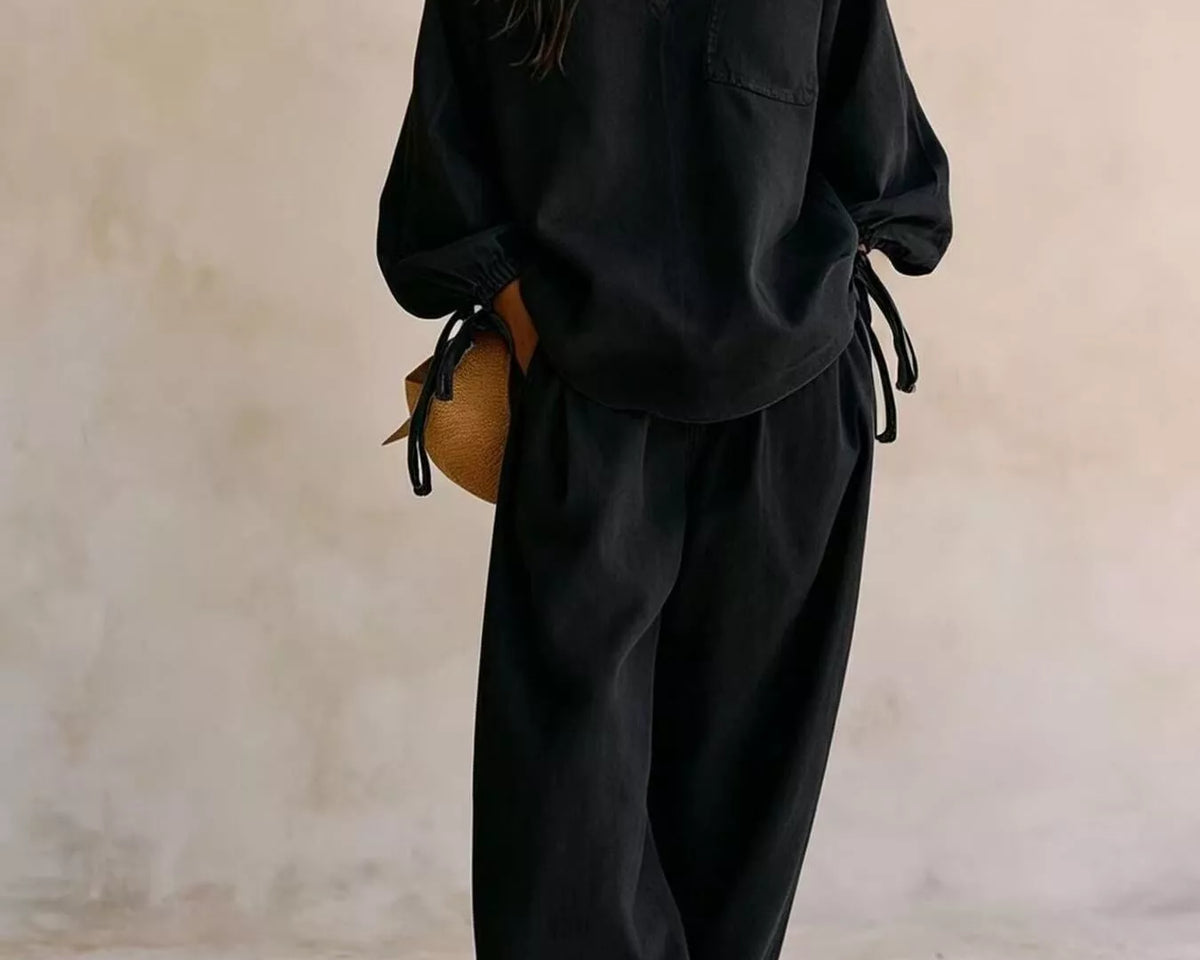 NEWYORKLISTED STYLE COLLECTION: A woman models a black two-piece lounge set consisting of a long-sleeve V-neck top with tie details at the cuffs and matching wide-leg pants, set against a textured beige wall.