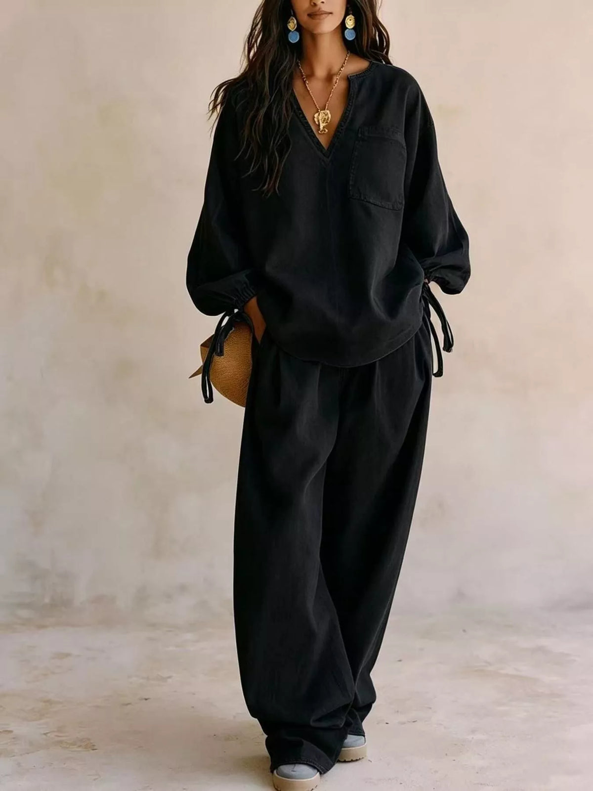 NEWYORKLISTED STYLE COLLECTION: A woman models a black two-piece lounge set consisting of a long-sleeve V-neck top with tie details at the cuffs and matching wide-leg pants, set against a textured beige wall.