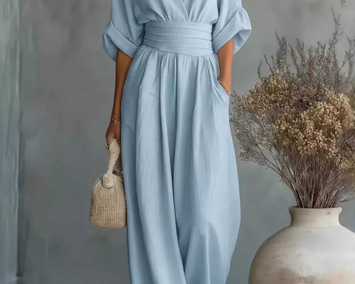 NEWYORKLISTED STYLE COLLECTION: Woman wearing a light blue dress standing next to a vase with dried flowers against a gray wall.