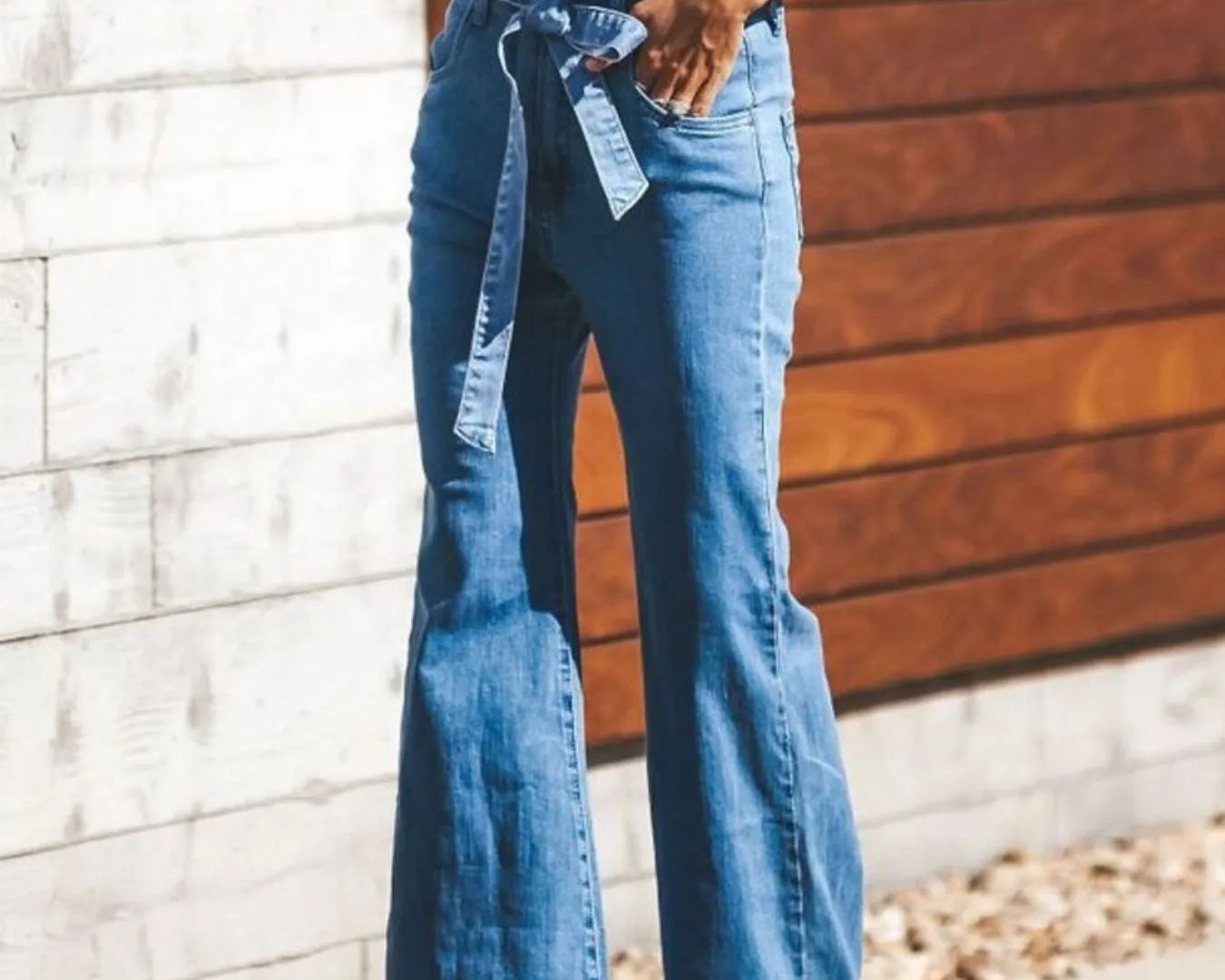 NEWYORKLISTED STYLE COLLECTION: Person wearing blue flared jeans with a belt, standing against a wooden and stone wall.