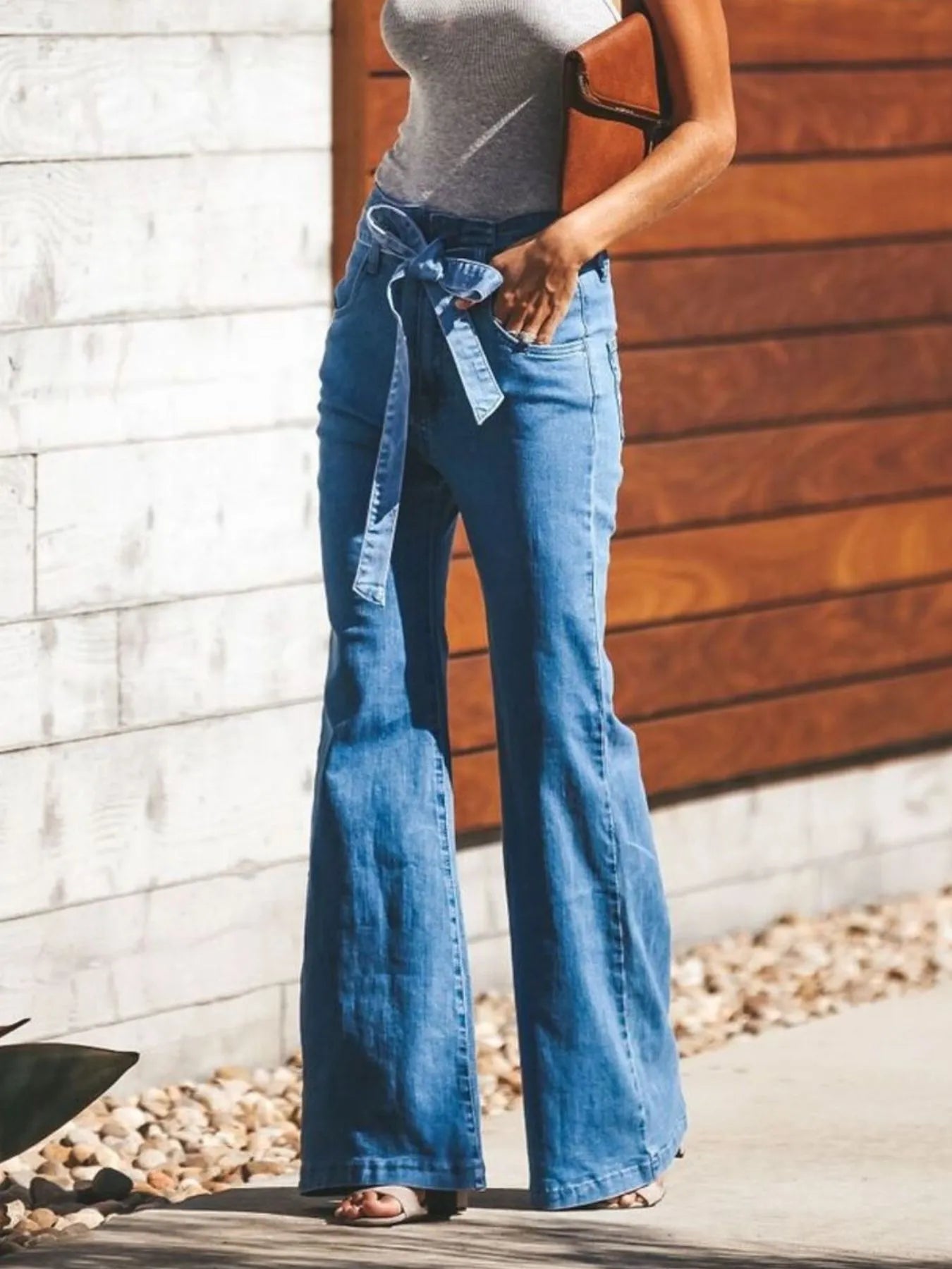 NEWYORKLISTED STYLE COLLECTION: Person wearing blue flared jeans with a belt, standing against a wooden and stone wall.