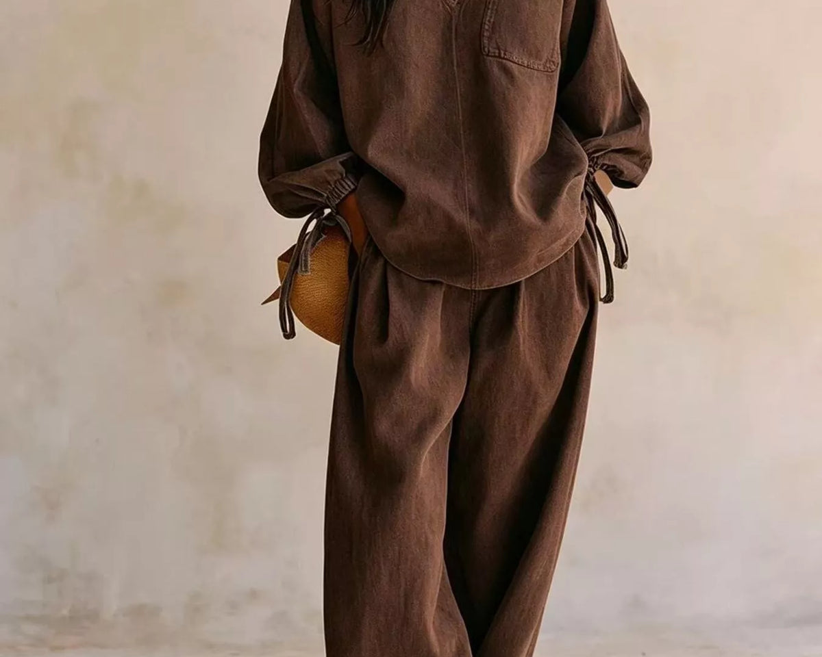 NEWYORKLISTED STYLE COLLECTION: A woman wearing a matching brown lounge set consisting of a long-sleeve V-neck top and wide-leg pants, styled with a small brown bag and gold jewelry against a textured beige wall.