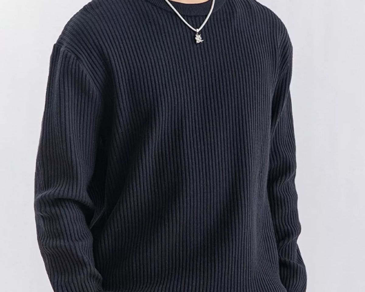 NEWYORKLISTED ATLAS MEN'S COLLECTION: A man wearing a dark navy ribbed knit crewneck sweater, paired with beige pants and a silver necklace, photographed against a light gray studio background.