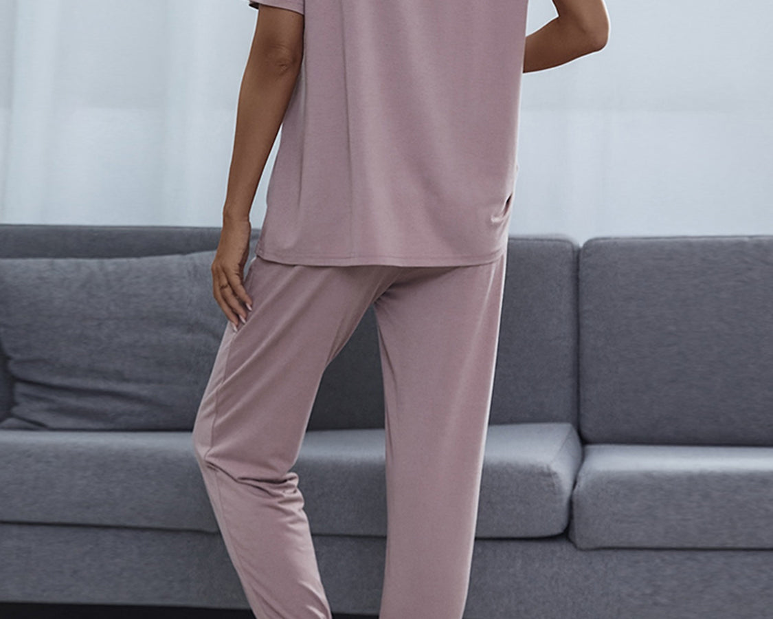 NEWYORKLISTED Intimate Apparel: Person wearing a mauve pajama set standing in a living room with a gray sofa.