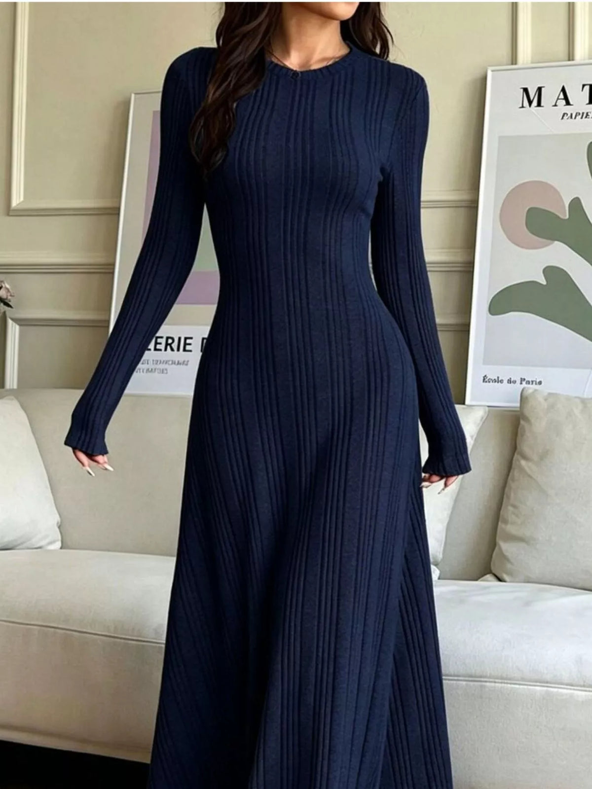 NEWYORKLISTED STYLE COLLECTION: A woman wearing a long-sleeve, ribbed knit maxi dress in dark navy blue, standing in a living room setting with a beige sofa and framed wall art in the background.
