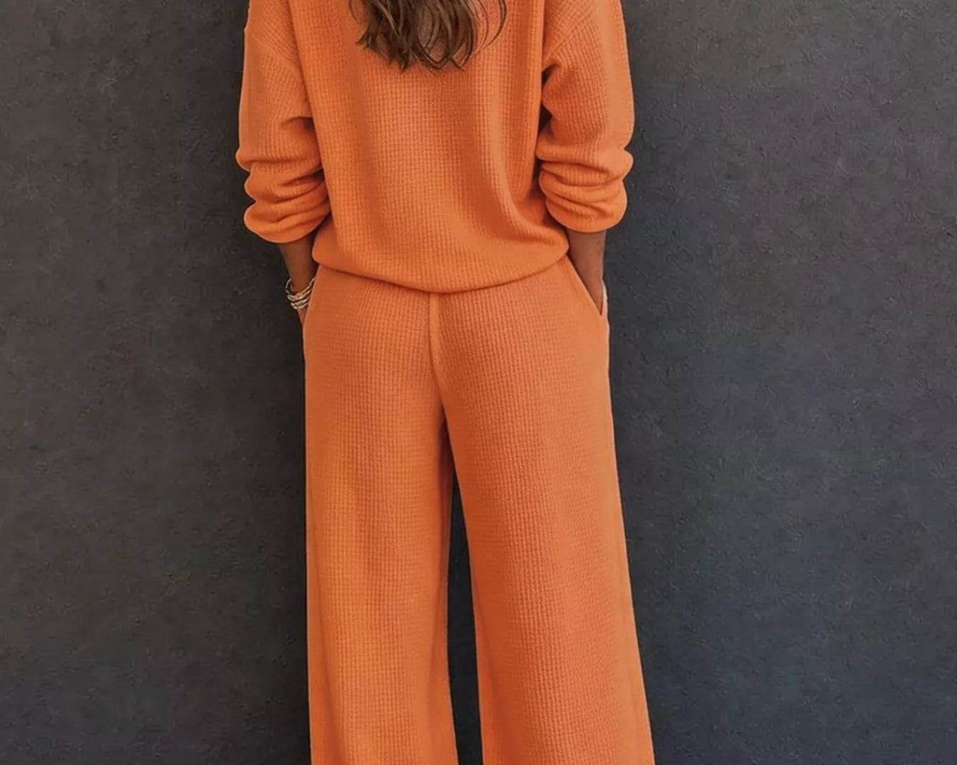 NEWYORKLISTED STYLE & COMFORT COLLECTION: A woman viewed from behind wearing a matching burnt orange knit lounge set consisting of a long-sleeve top and wide-leg pants, standing against a dark gray textured wall.