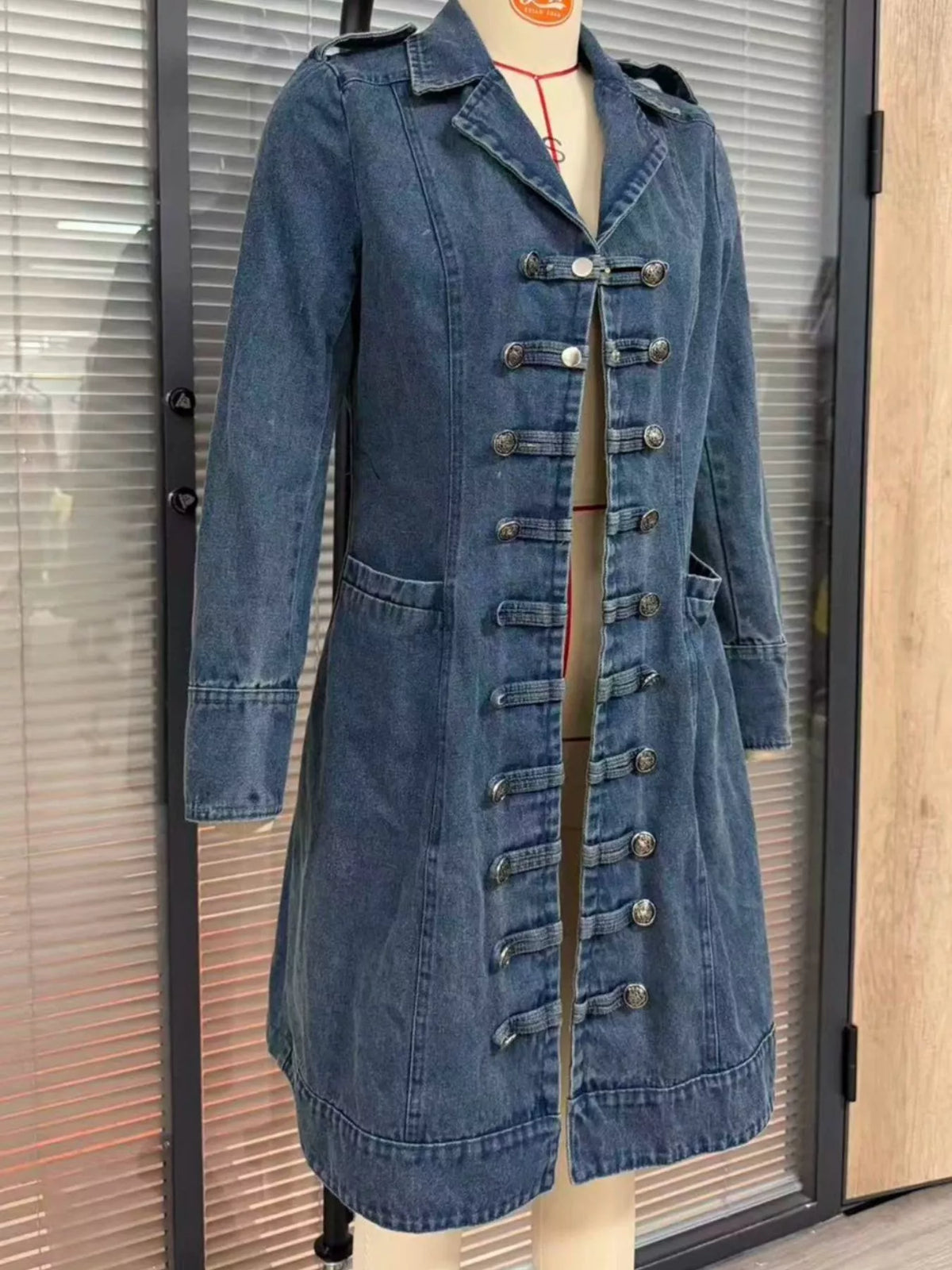 NEWYORKLISTED STYLE COLLECTION – Women's long blue denim military-inspired coat featuring a notched lapel collar, decorative silver-tone buttons, double-breasted styling, long sleeves, front pockets, and a tailored silhouette with a flared hem.