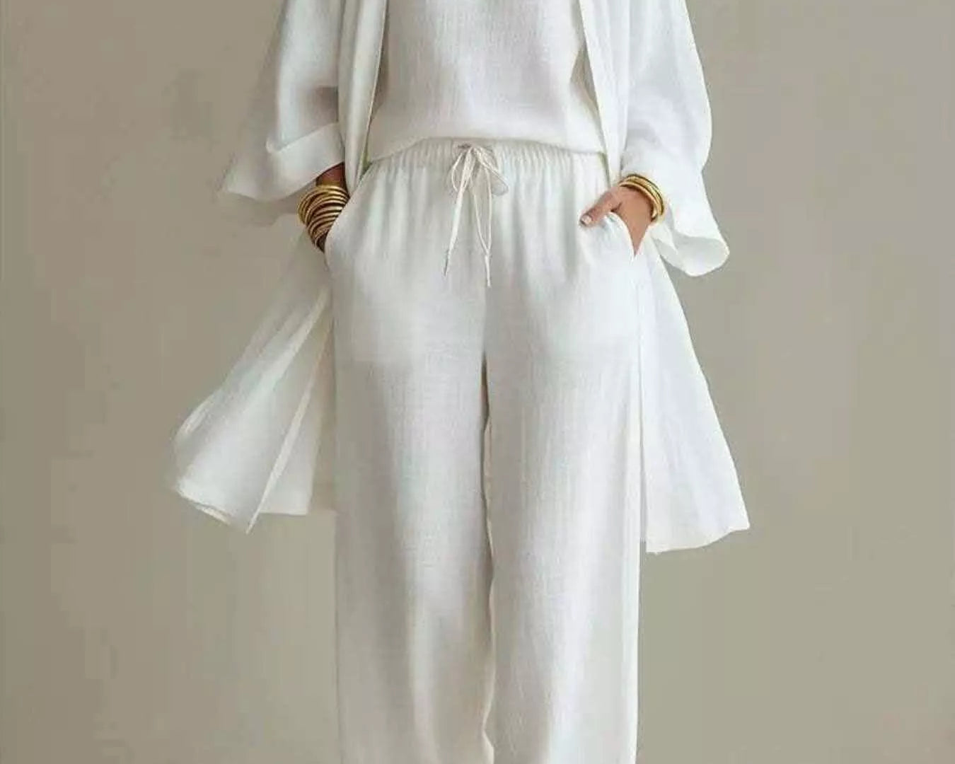 NEWYORKLISTED STYLE COLLECTION: A woman modeling a white three-piece linen outfit consisting of a V-neck top, matching wide-leg pants with a drawstring, and a lightweight open-front cardigan. She is accessorized with layered gold necklaces and bracelets, standing against a neutral beige studio background.
