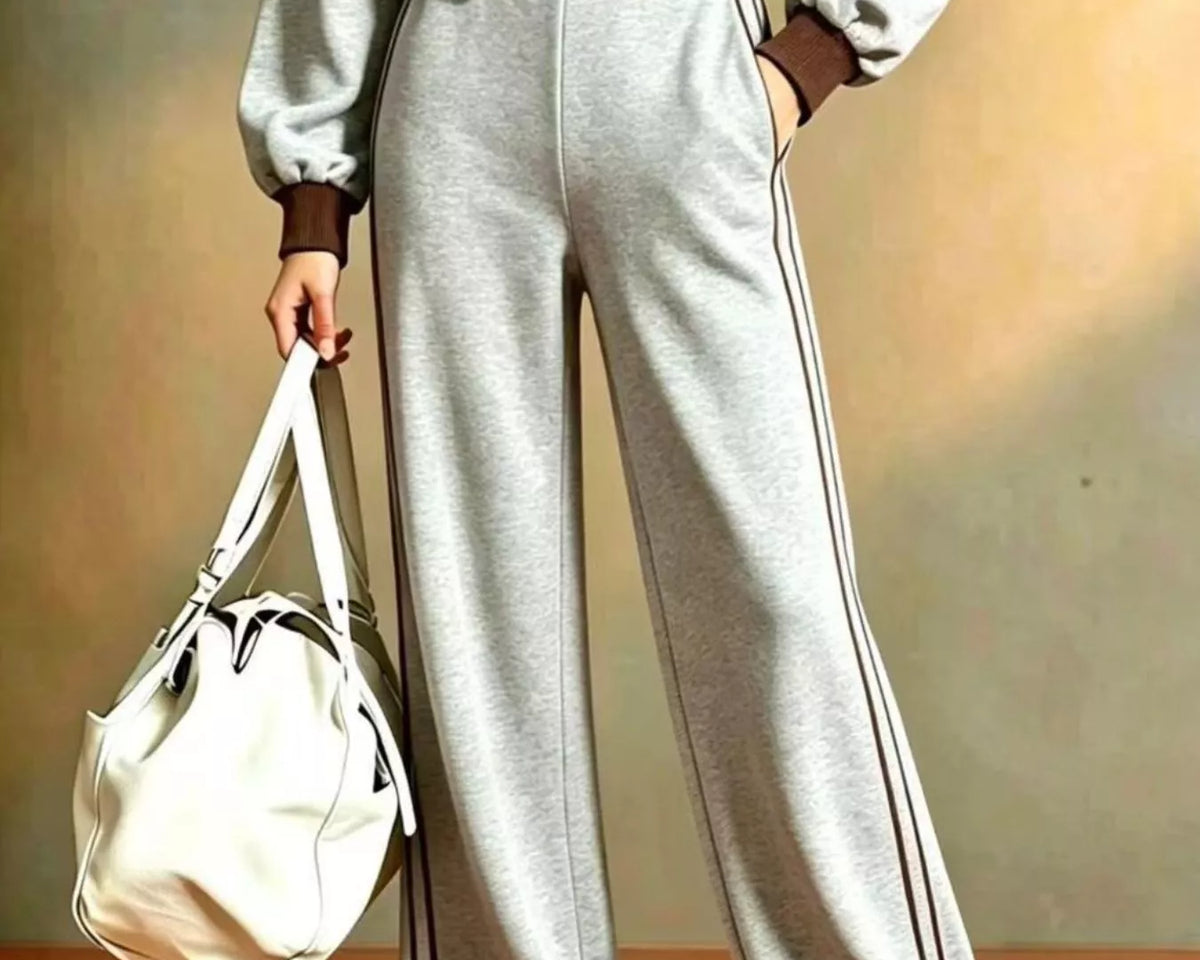 NEWYORKLISTED STYLE COLLECTION: A person wearing a light gray athletic tracksuit with brown trim, including a cropped zip-up jacket and wide-leg pants, holding a white shoulder bag against a warm beige studio background.