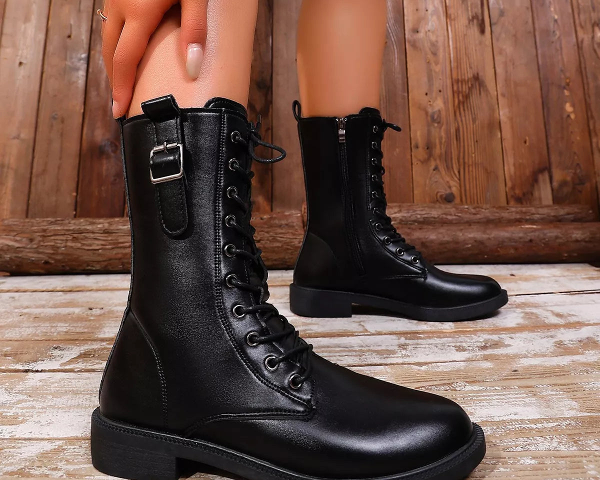 NEWYORKLISTED SHOE COLLECTION: A pair of black leather lace-up ankle boots with side buckle details, shown on a model's feet against a rustic wooden plank background.