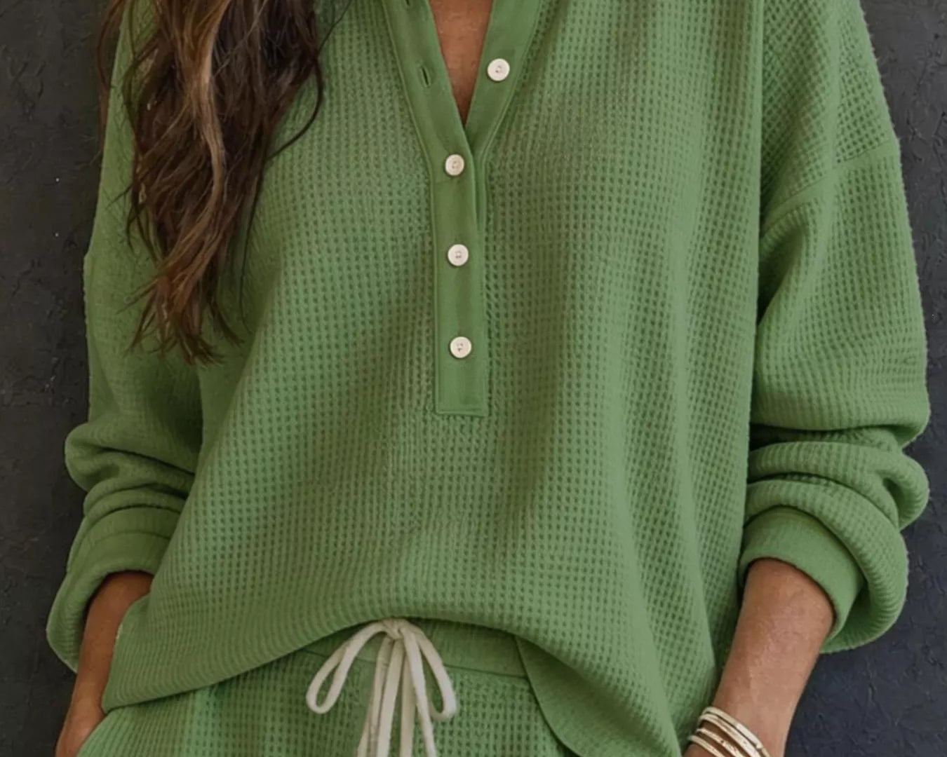 NEWYORKLISTED STYLE & COMFORT COLLECTION: A woman wearing a sage green waffle-knit long-sleeve henley top with white buttons and matching green drawstring pants, standing against a dark textured wall.