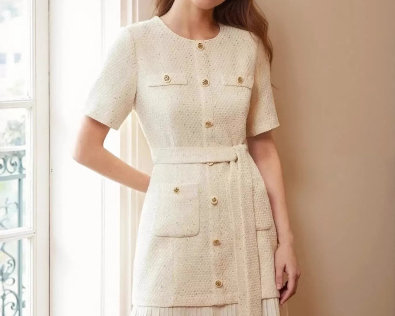 NEWYORKLISTED STYLE COLLECTION: Woman wearing a cream-colored dress with buttons standing in a room with a window.