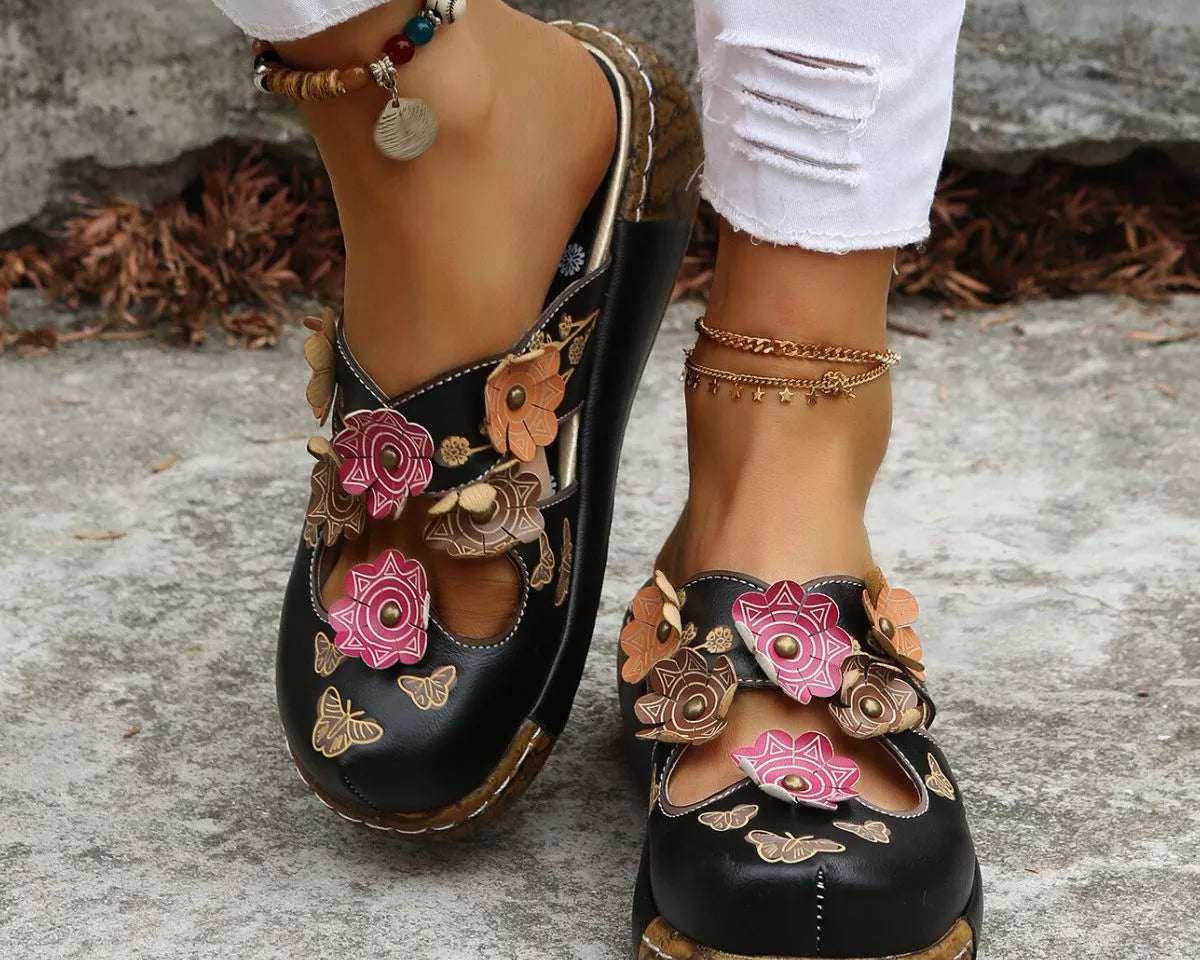 NEWYORKLISTED SHOE COLLECTION: Black floral sandals worn with white pants on a stone surface
